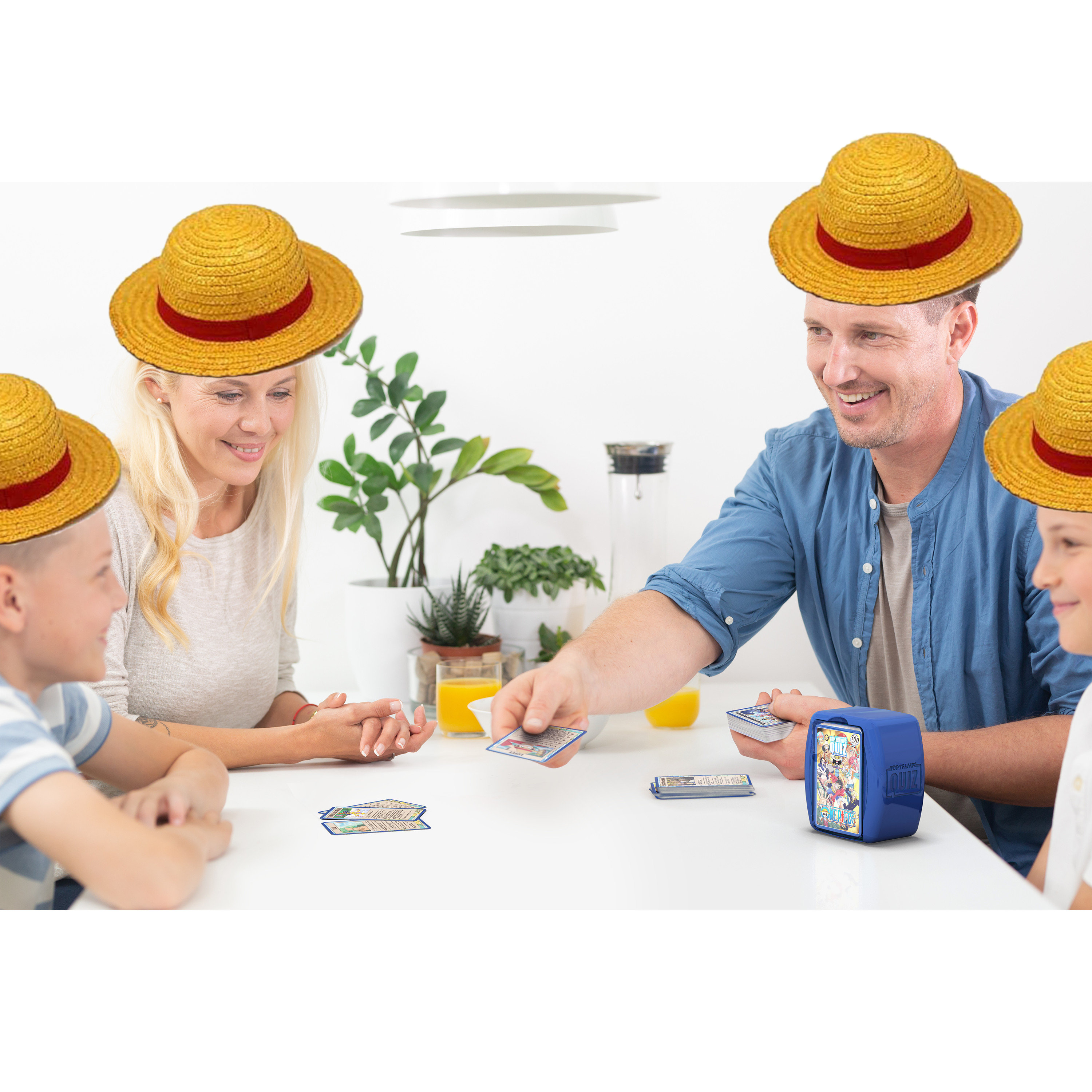Familie spielt Kartenspiel. Alle tragen Strohhüte. Karten und blaue Box auf weißem Tisch.