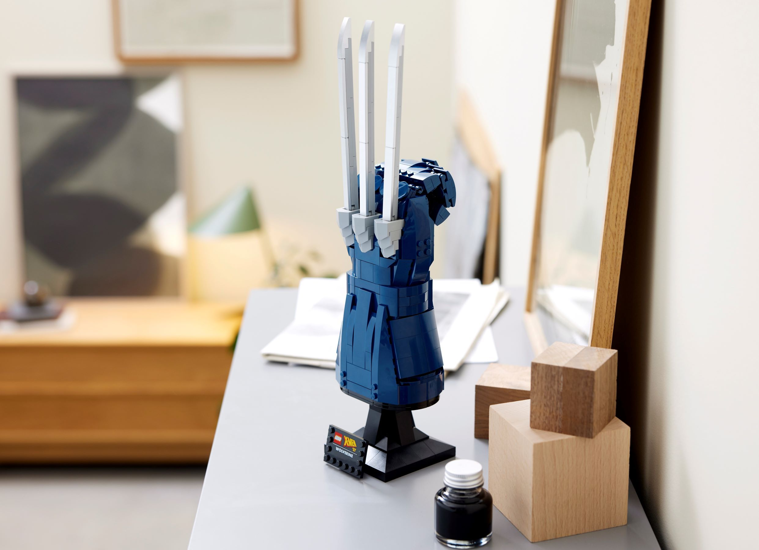 A LEGO Wolverine hand with claws, standing on a black base with a small label, near wooden blocks and ink.