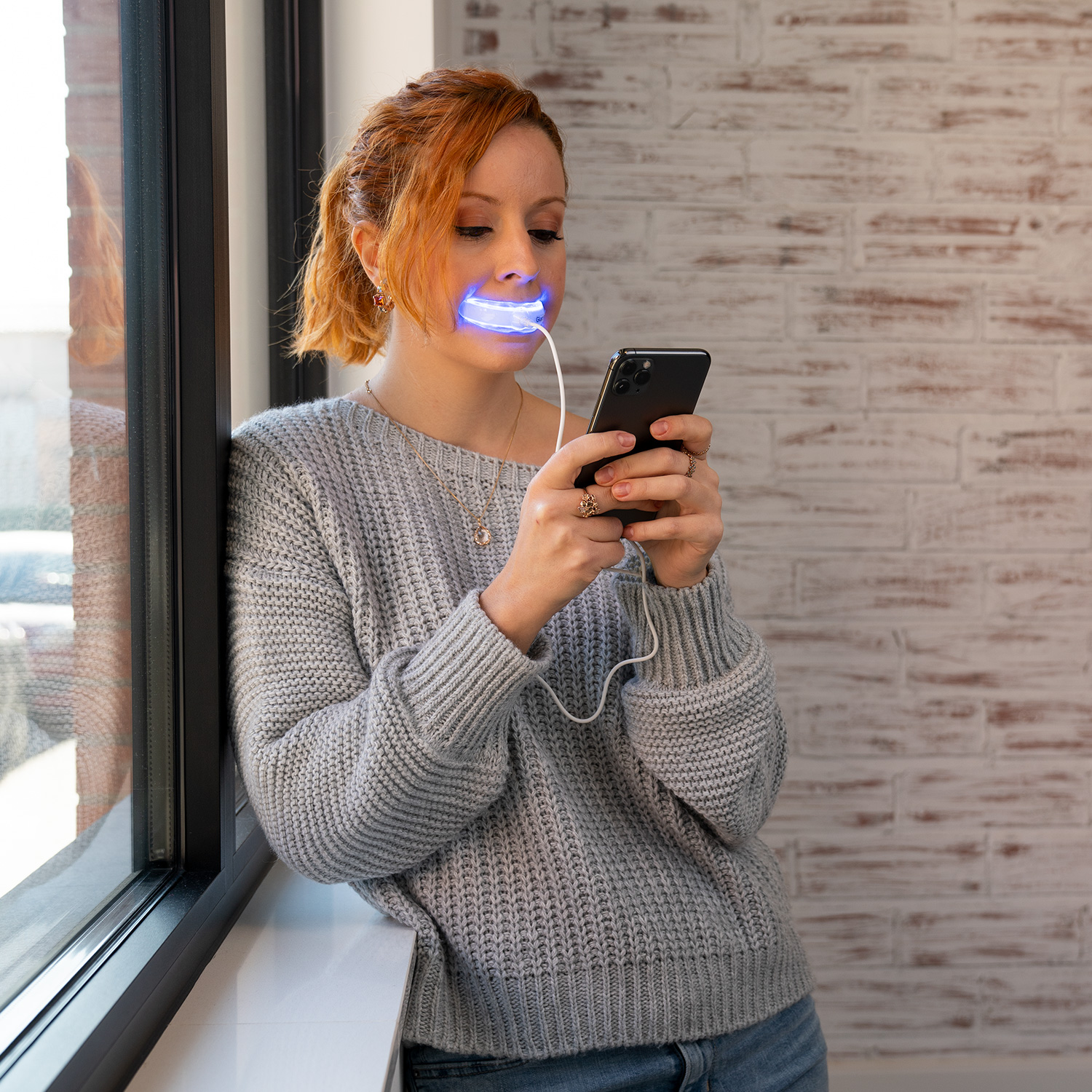 Mujer usando un teléfono, usando un blanqueador dental con luz, cerca de una ventana.