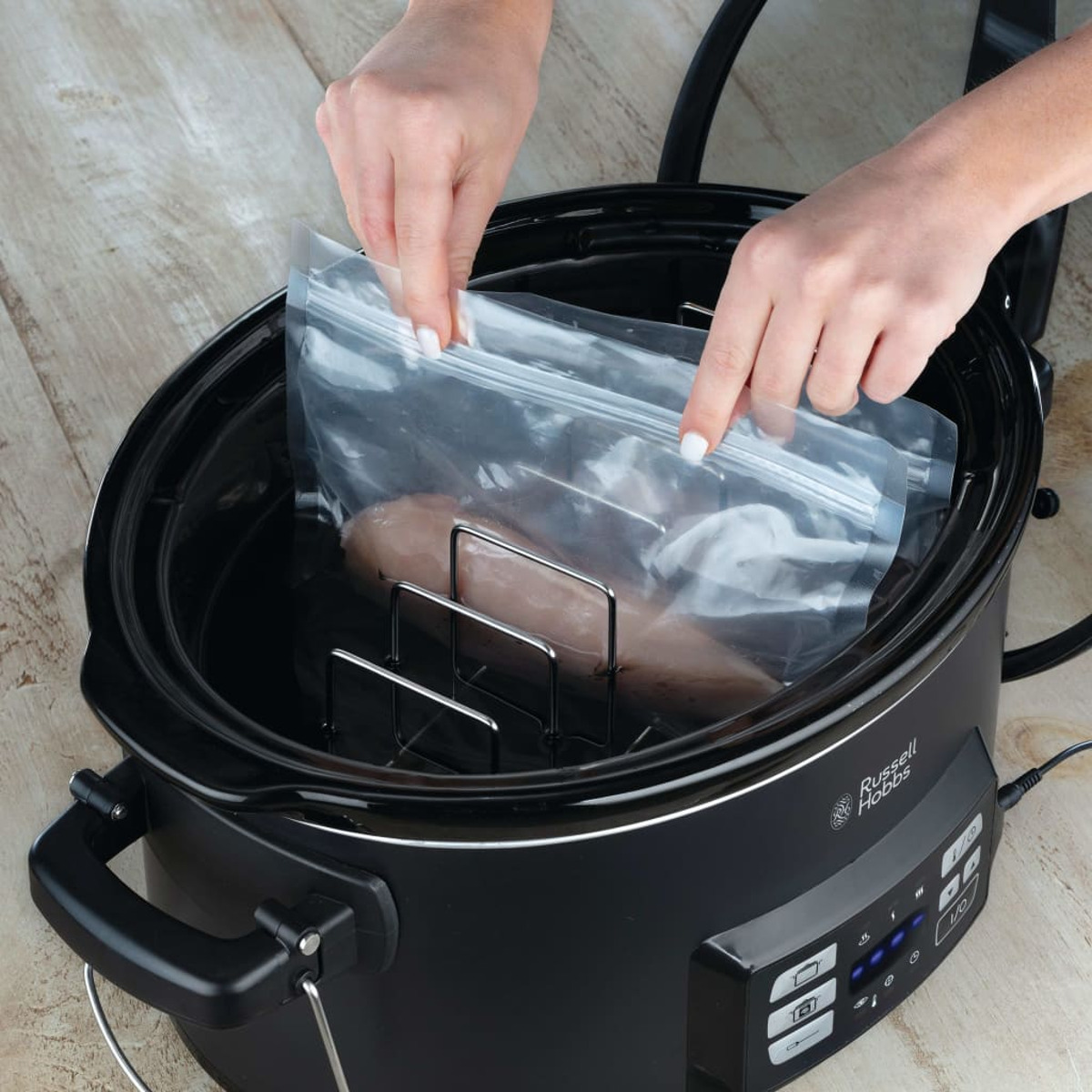 Hände legen einen Beutel in einen schwarzen Slow Cooker. Ein Stück Fleisch ist im Beutel.