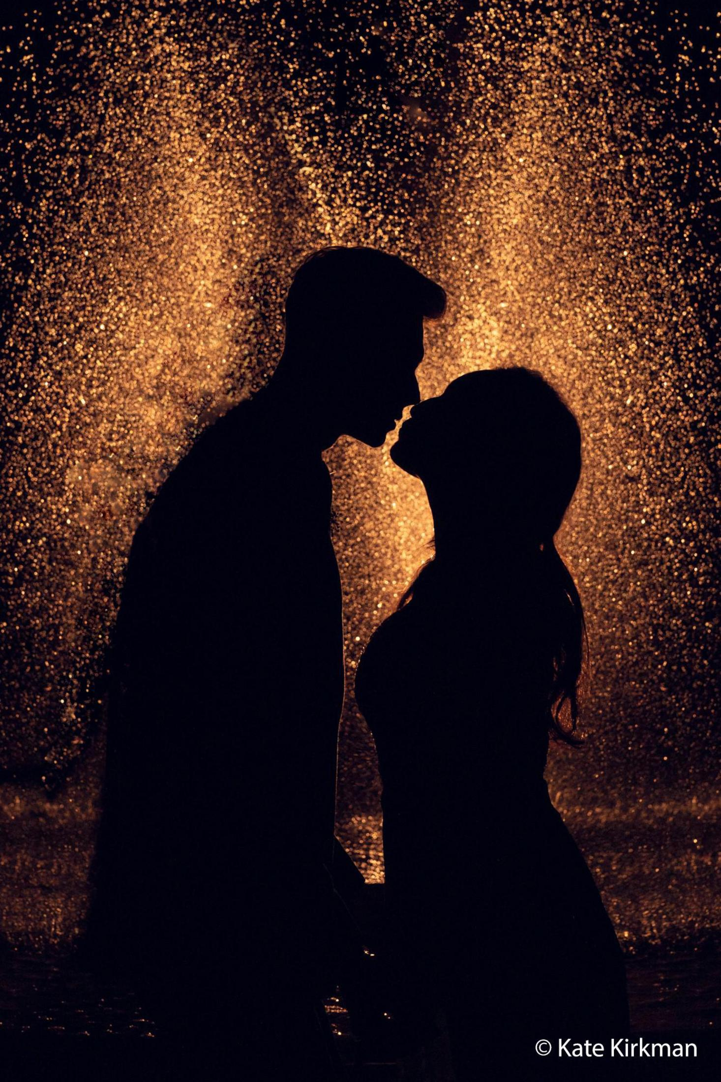 Silhouettenpaar küsst sich vor goldenem Wasserfontäne. Dunkler Hintergrund mit Wasserspray.