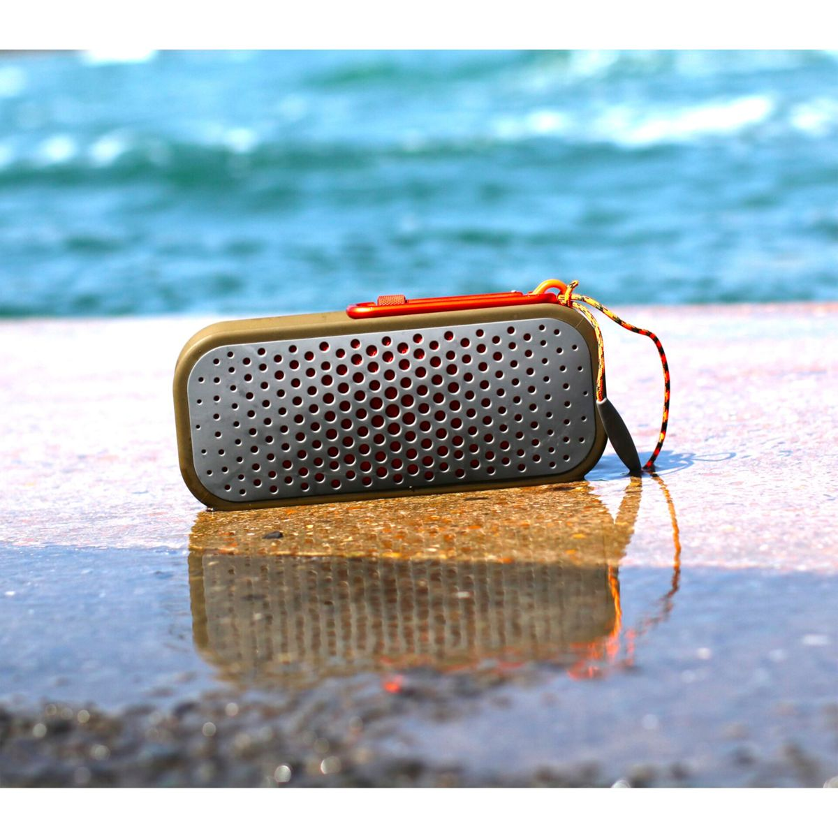 Un altoparlante portatile, verde oliva e arancione, su una superficie bagnata, con una spiaggia sullo sfondo.