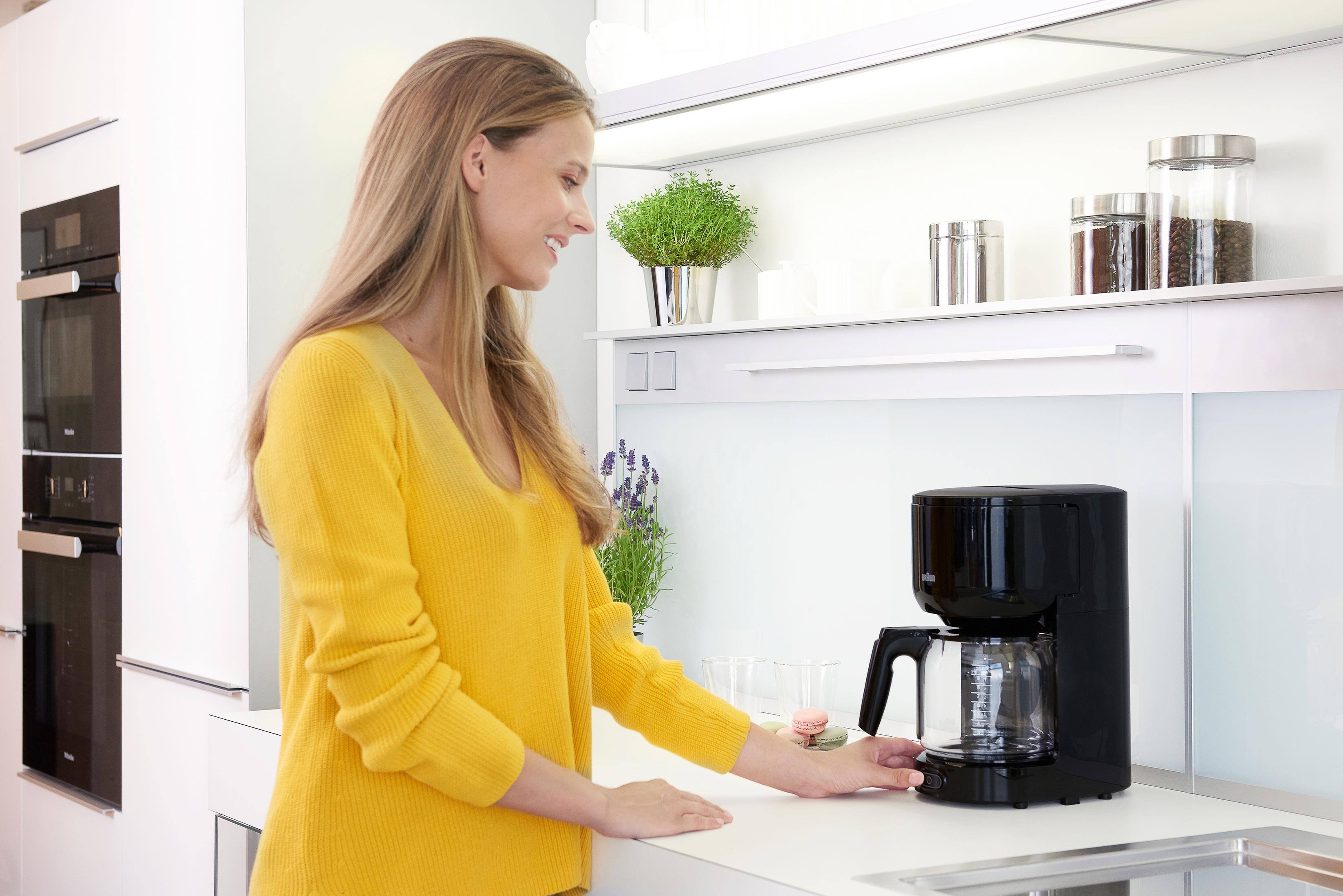 Kobieta w żółtym swetrze obsługuje czarny ekspres do kawy na białym blacie.