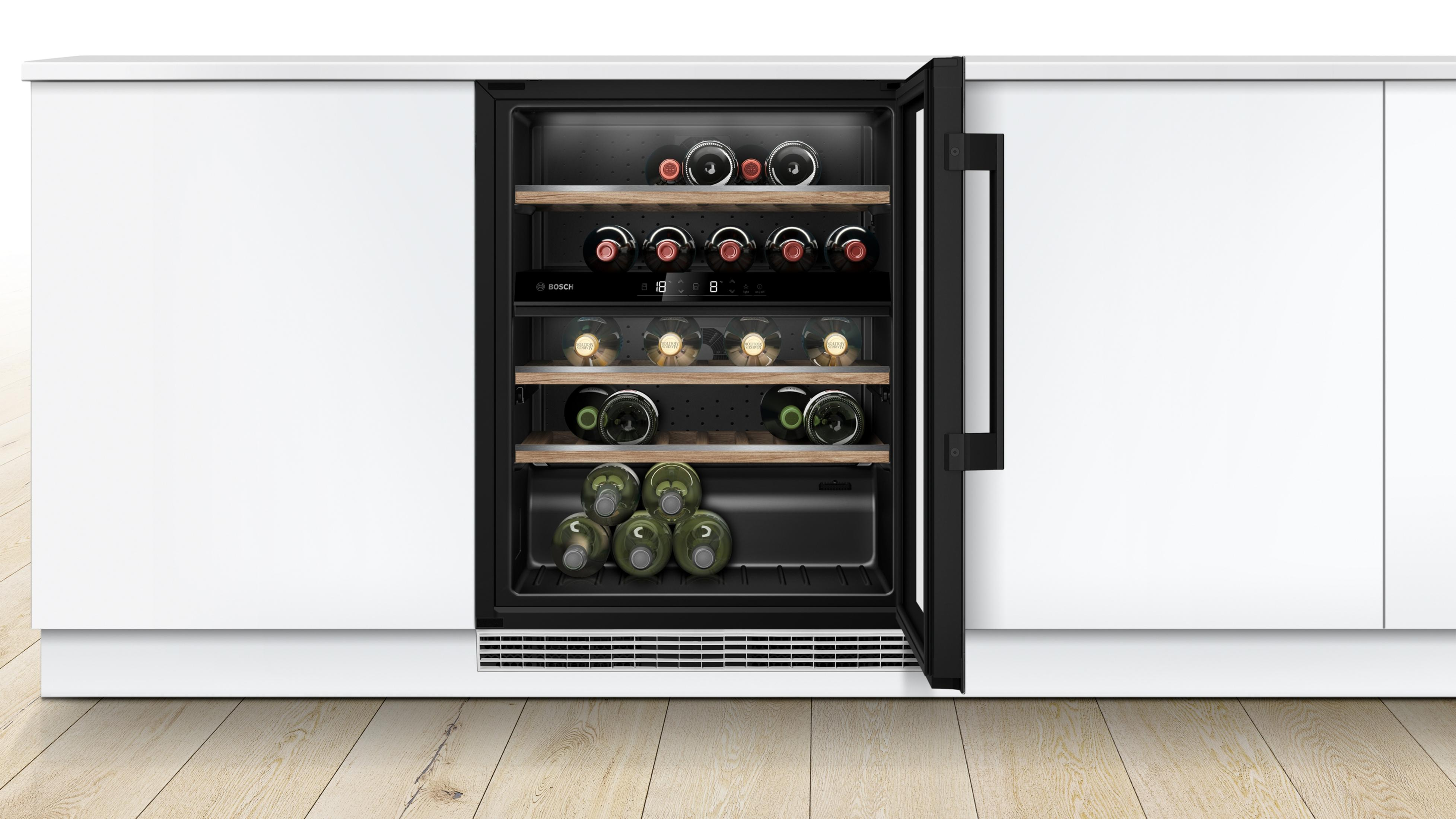 Enfriador de vino empotrado con puerta abierta, mostrando botellas. Armarios blancos, suelo madera.