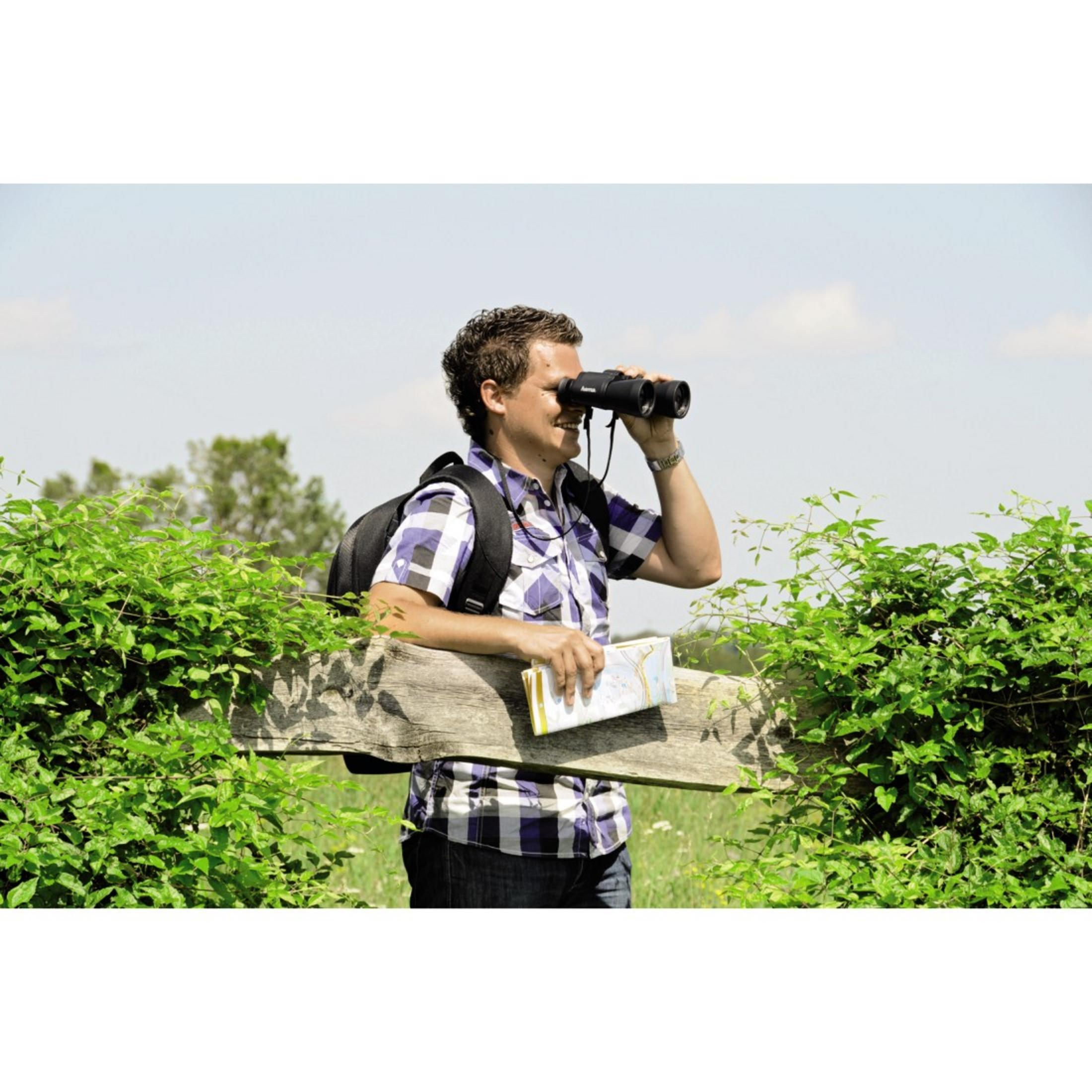 Mężczyzna w koszuli w kratę patrzy przez lornetkę, mapa w ręku, zielone krzaki, słoneczny dzień.