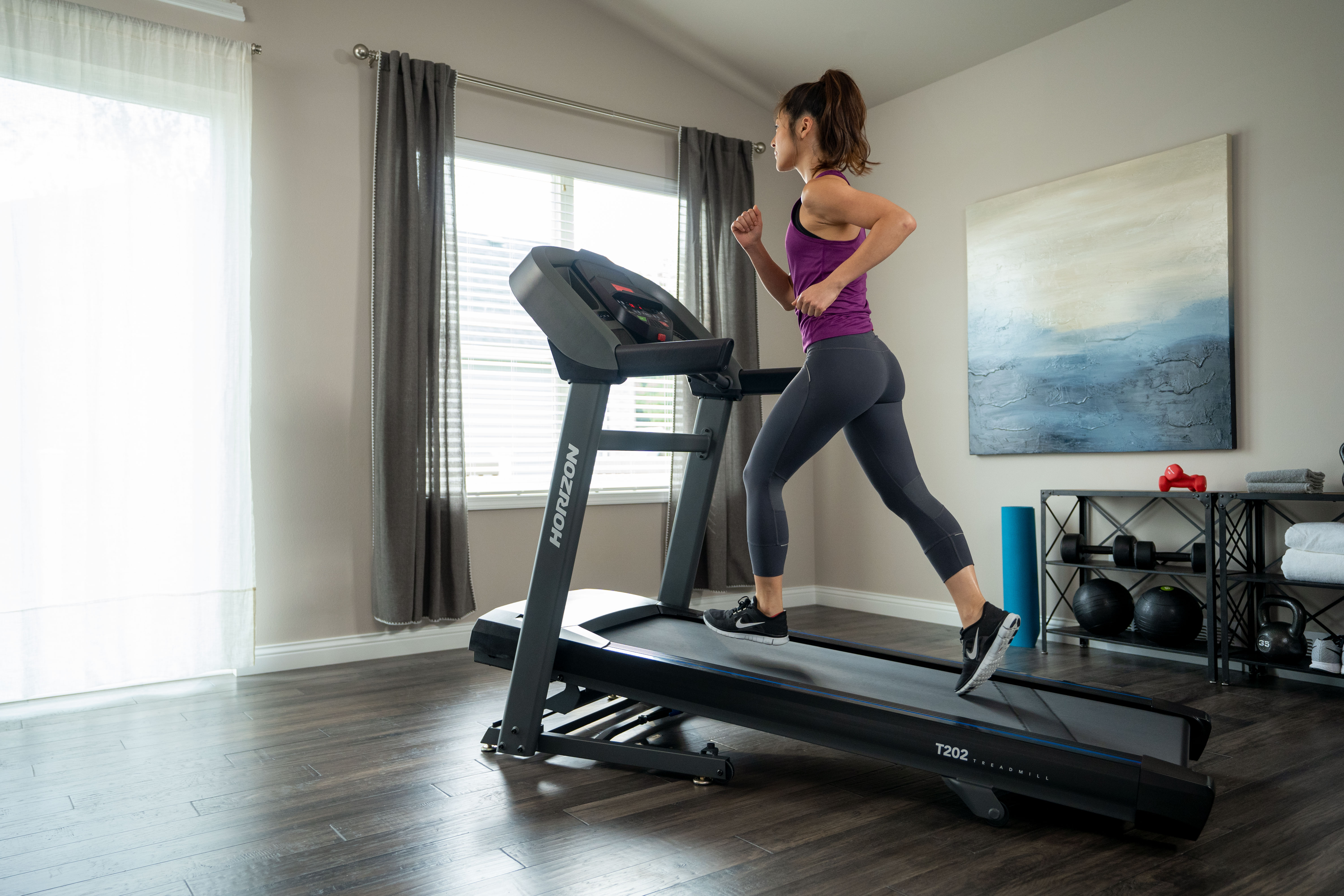 Kobieta biegnąca na bieżni w pomieszczeniu. Pokój ma drewniane podłogi i okno z żaluzjami.