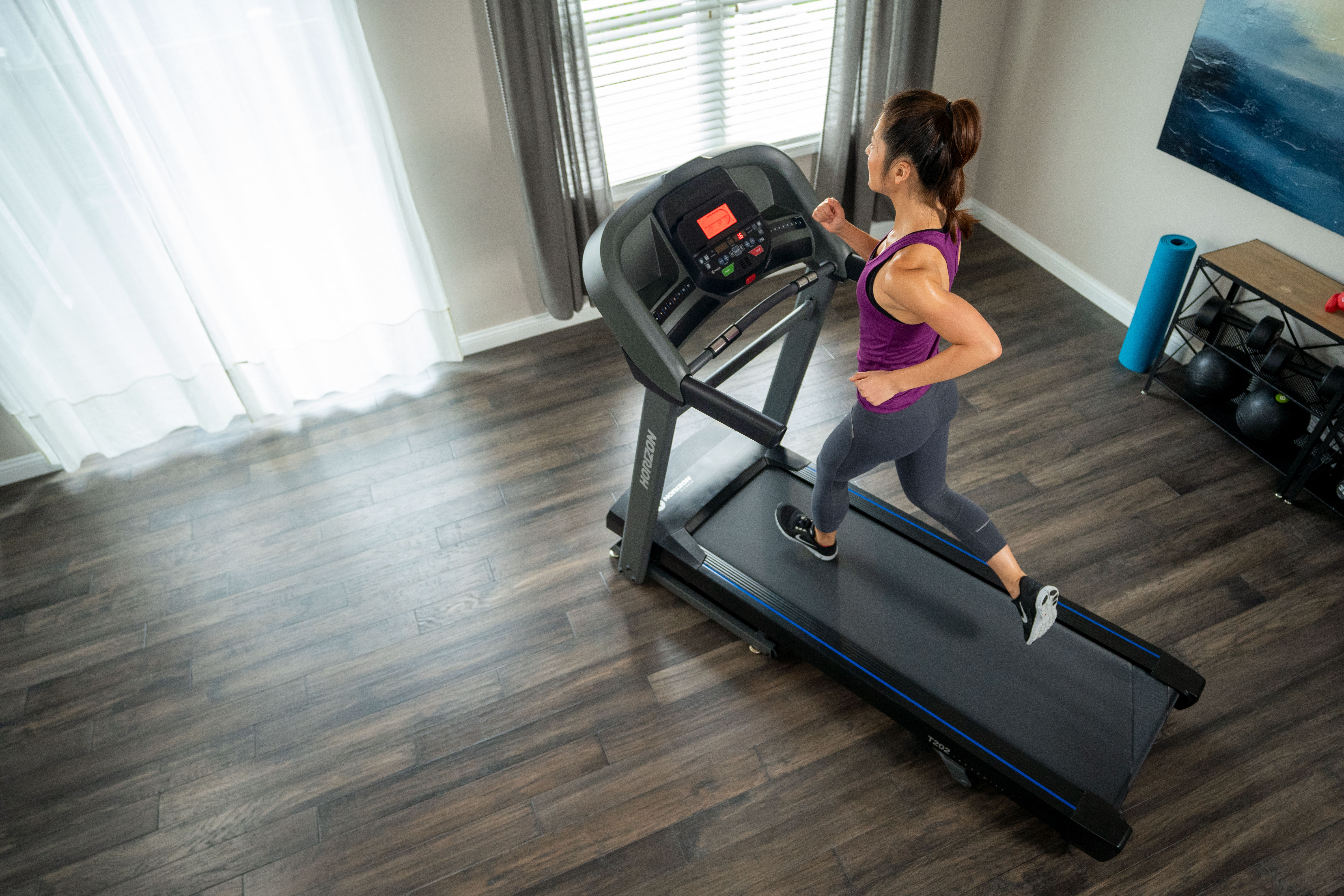 Kobieta biegnie na bieżni. Pokój ma okno, niebieską matę i półkę.