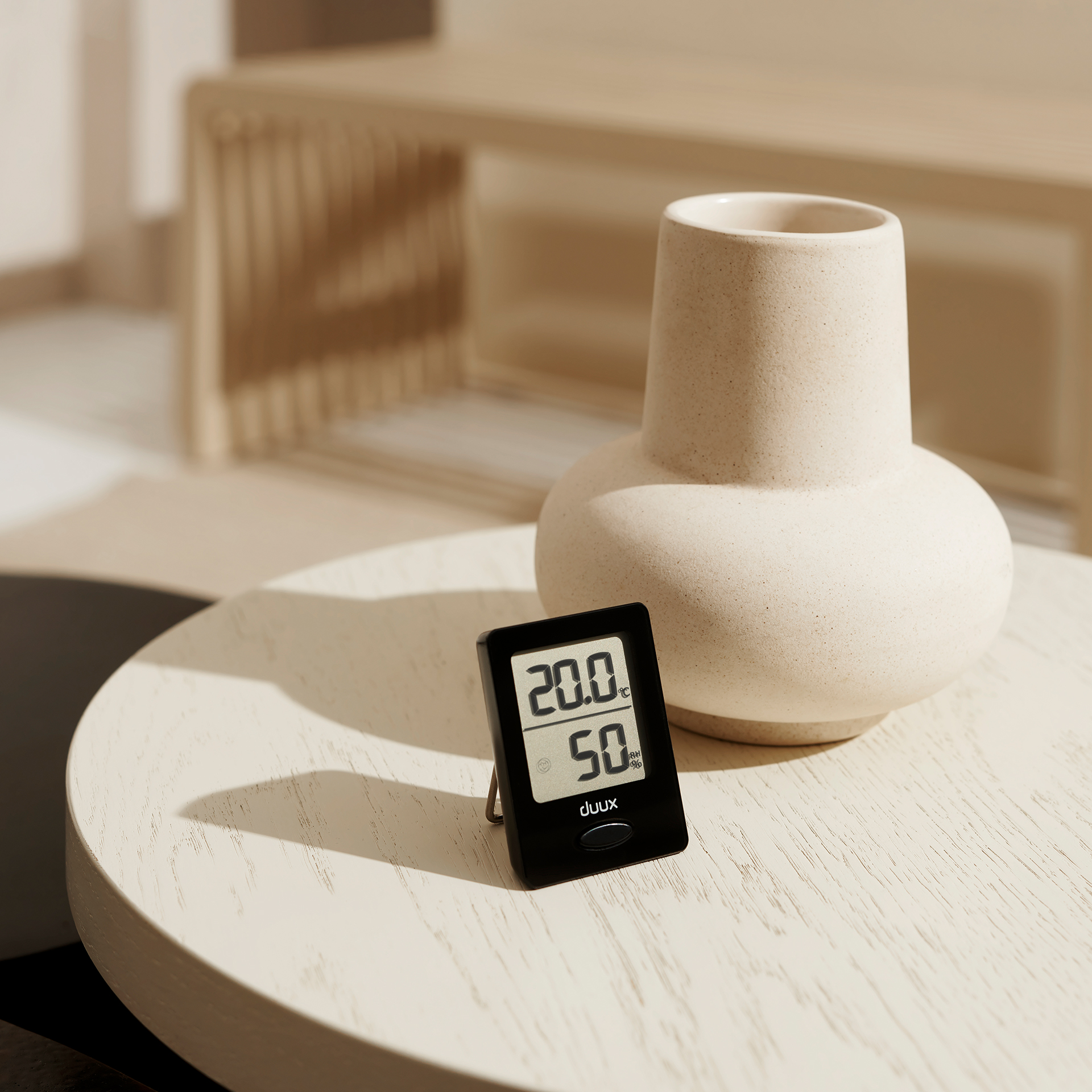 Un termómetro negro muestra 20,0 y 50 en una mesa de madera con un jarrón.