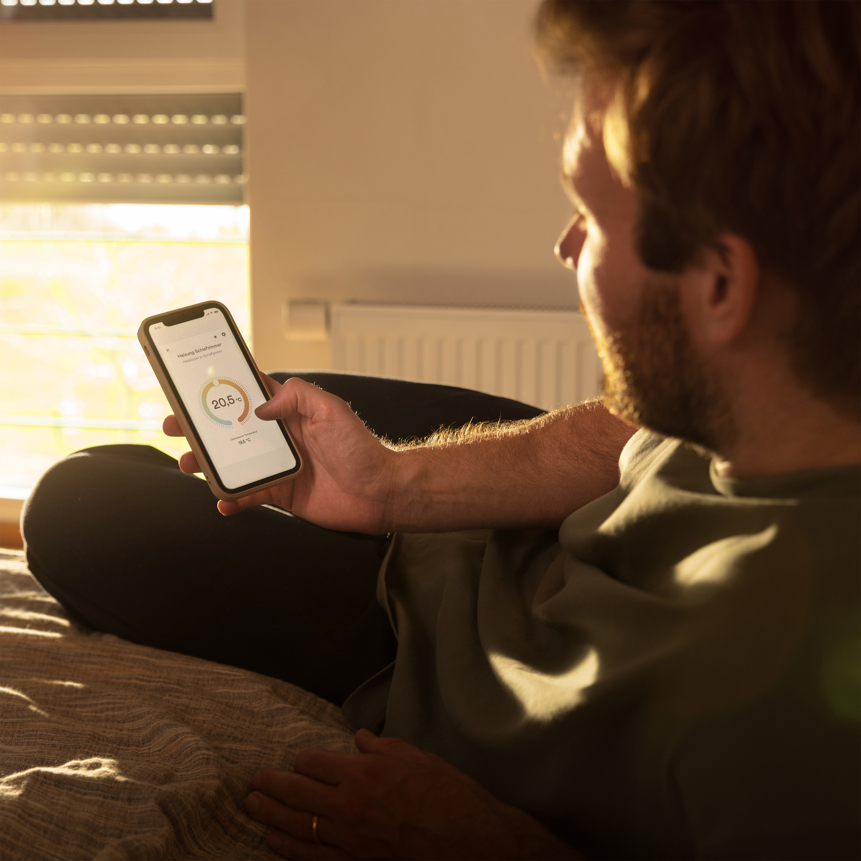 Mann auf einem Bett, der ein Telefon mit einer App hält. Sonnenbeschienenes Fenster im Hintergrund.