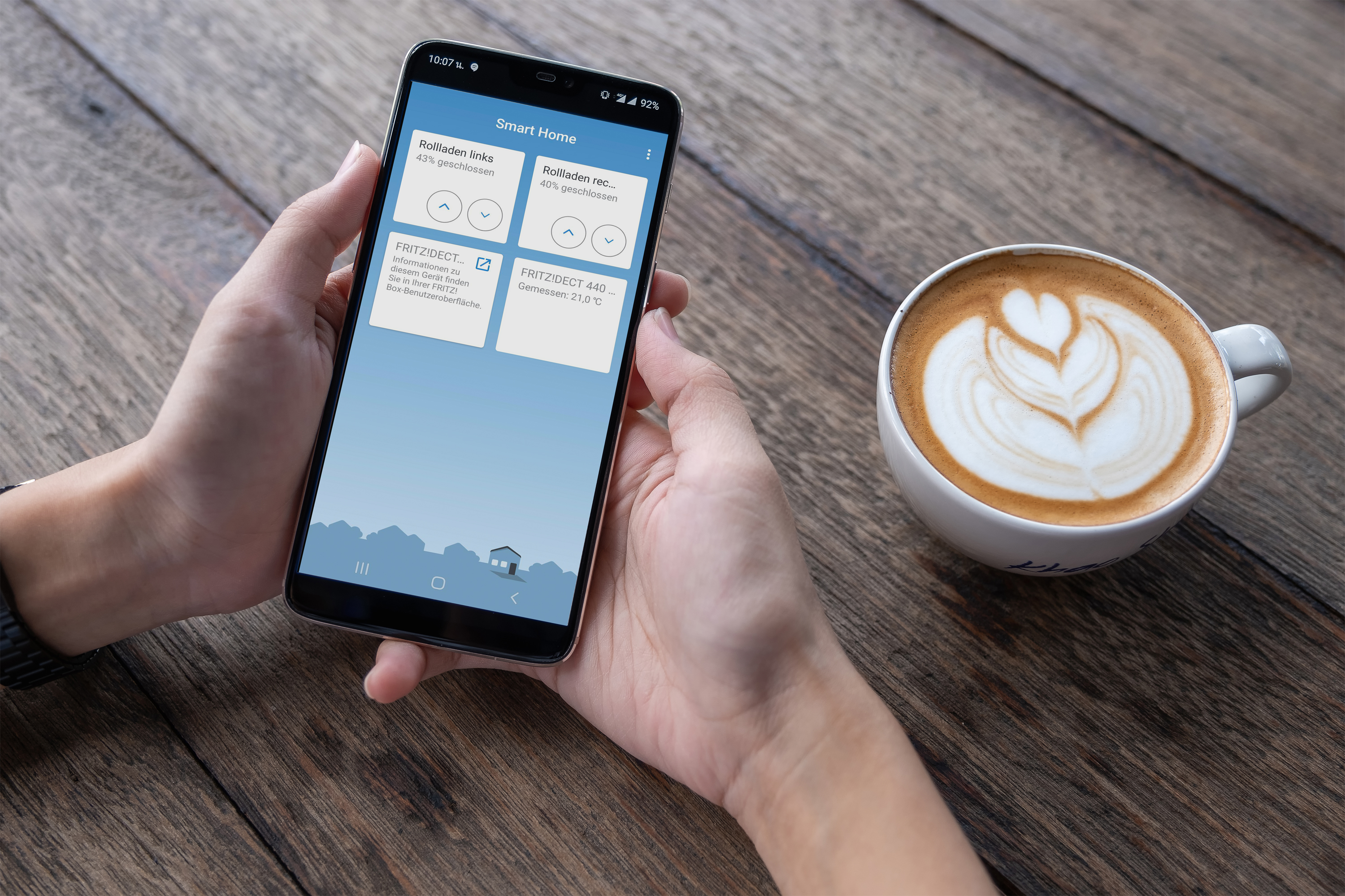 Hände halten Smartphone mit Smart-Home-App neben Kaffee mit Latte-Art.