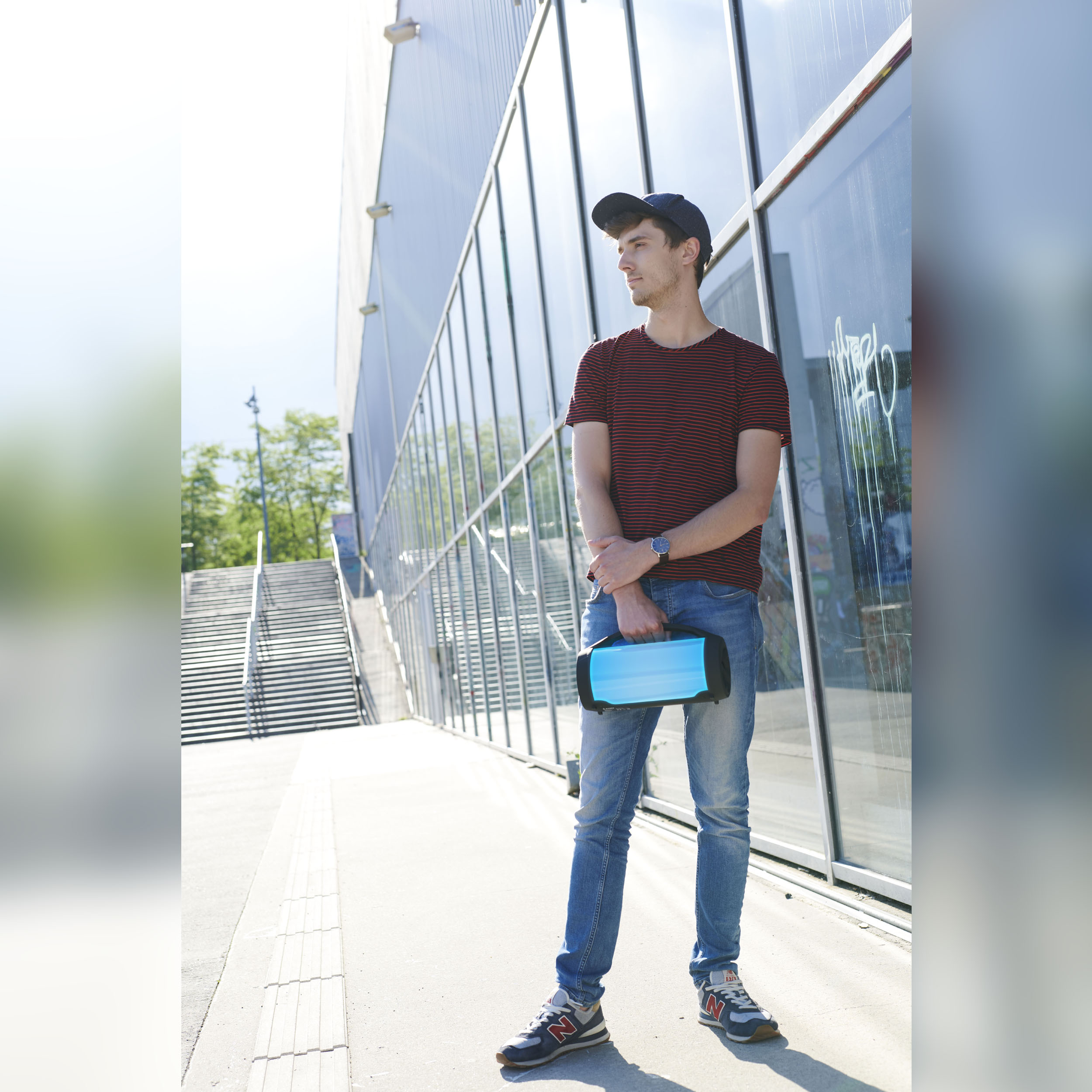 Mężczyzna w koszuli w paski i dżinsach stoi z niebieskim głośnikiem przed szklanym budynkiem.