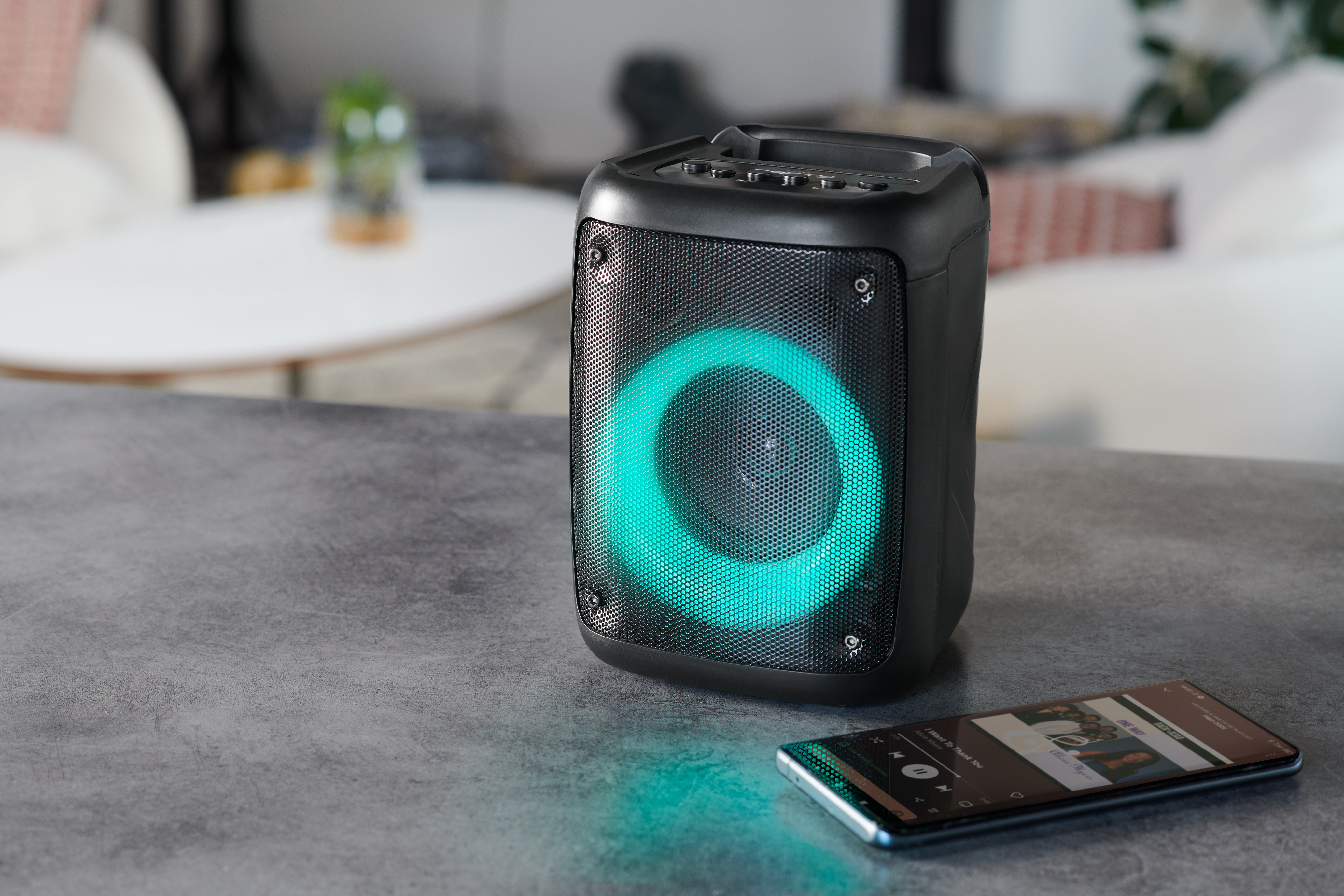 Czarny głośnik ze świecącym zielonym światłem. Obok na stole leży smartfon.