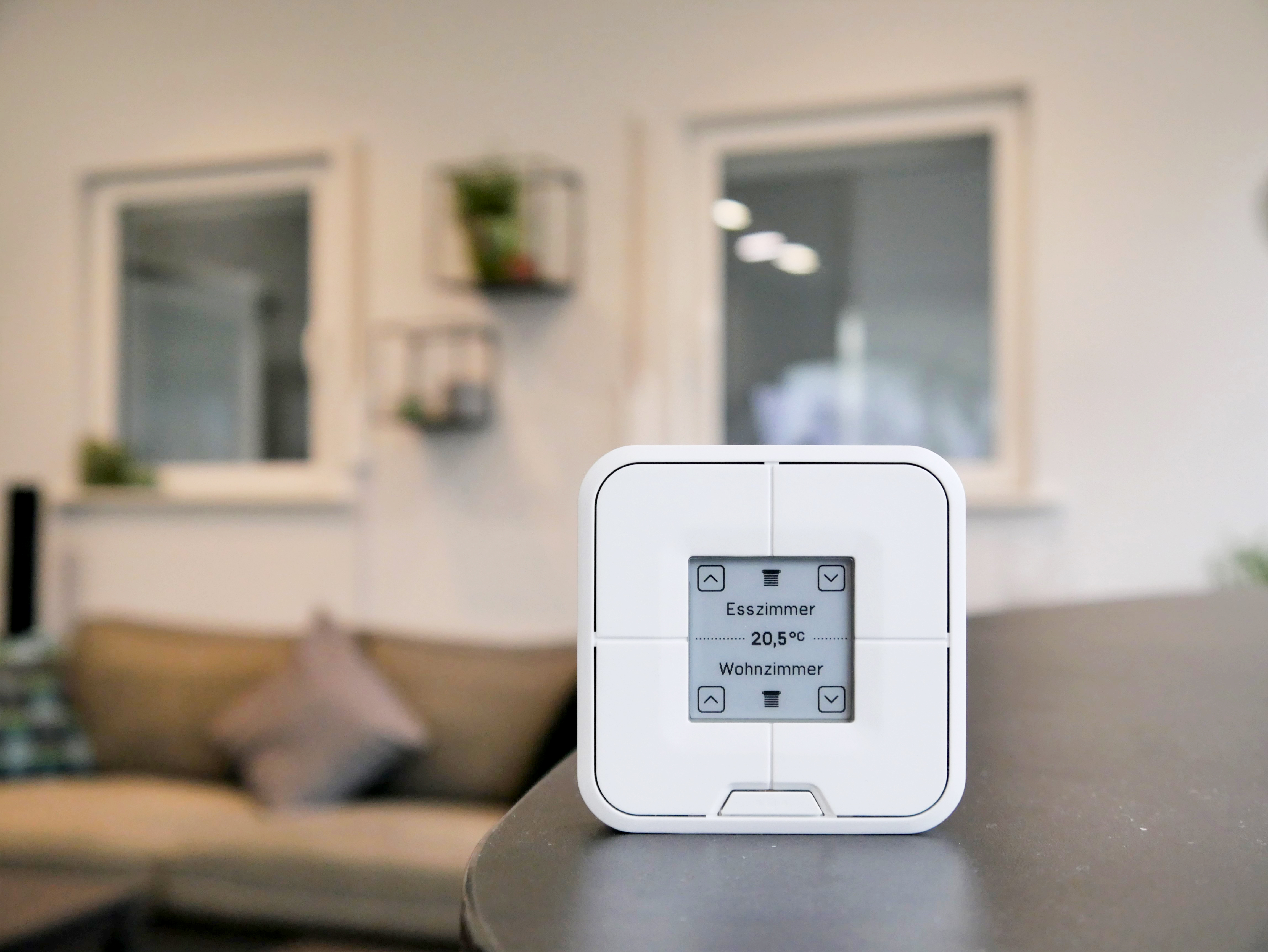 Ein weißer intelligenter Thermostat, der Temperatur und Raumnamen in einer häuslichen Umgebung anzeigt.