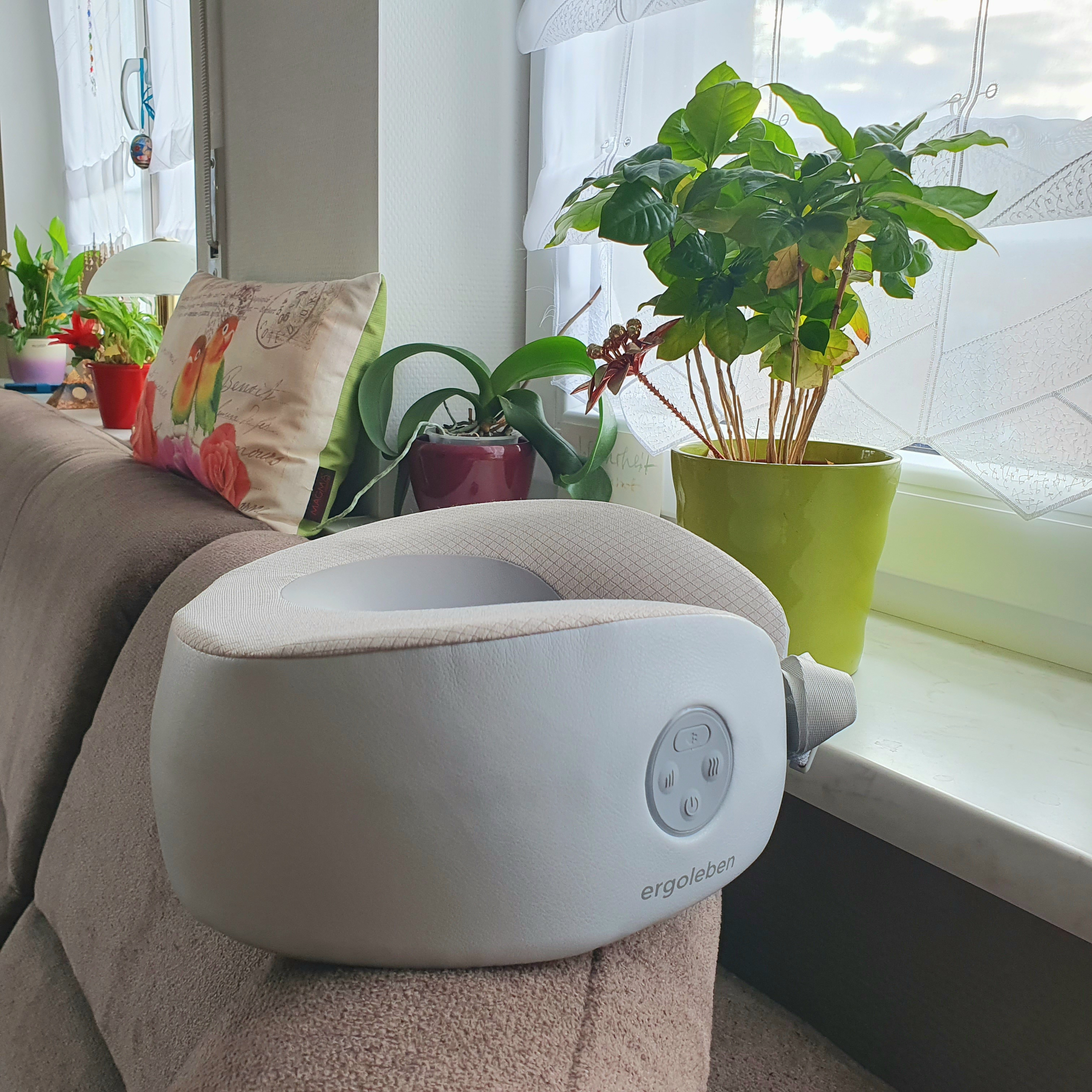 Weißer Nackenmassagegerät auf Sofa, nahe Pflanzen und Fenster.