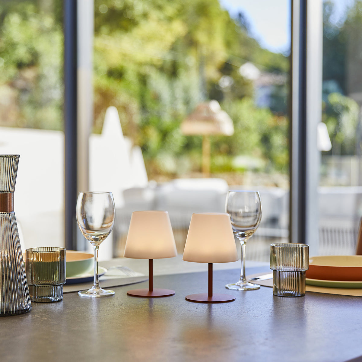Zwei Lampen auf einem Tisch, Weingläser und eine Karaffe. Im Hintergrund ist ein Garten zu sehen.