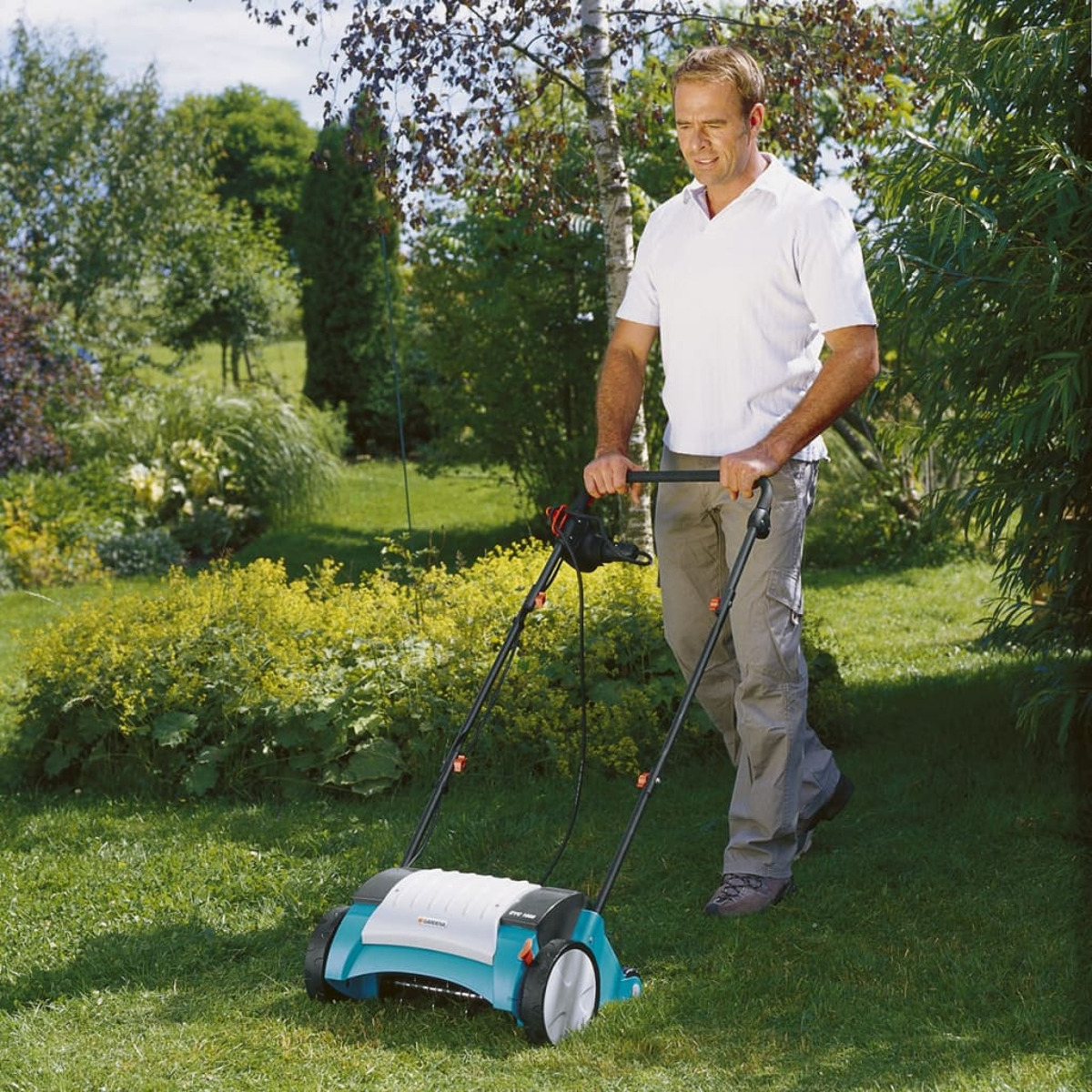 Un uomo che arieggia un prato con un arieggiatore blu e bianco, prato verde e alberi.