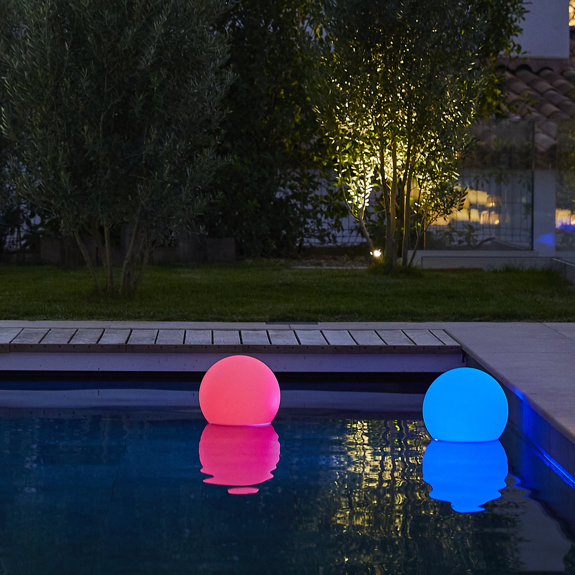 Zwei leuchtende Kugeln, eine rosa, eine blaue, schwimmen in einem Pool. Bäume und ein Haus sind im Hintergrund.