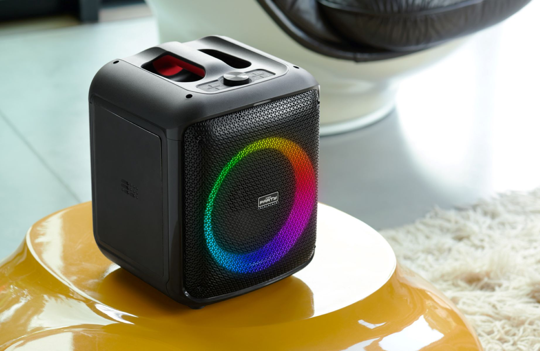 Czarny głośnik z kolorowym pierścieniem świetlnym na żółtym stole, białe tło.