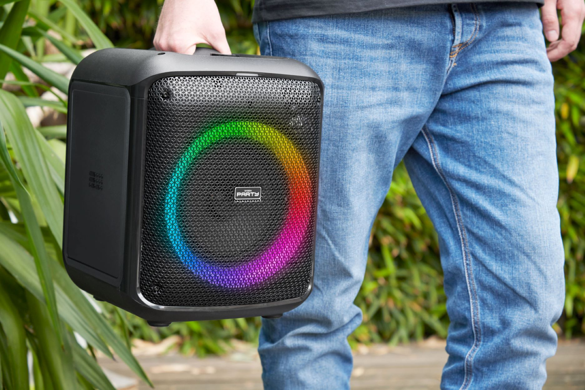 Persona sostiene altavoz negro con anillo brillante de colores. Plantas verdes en el fondo.