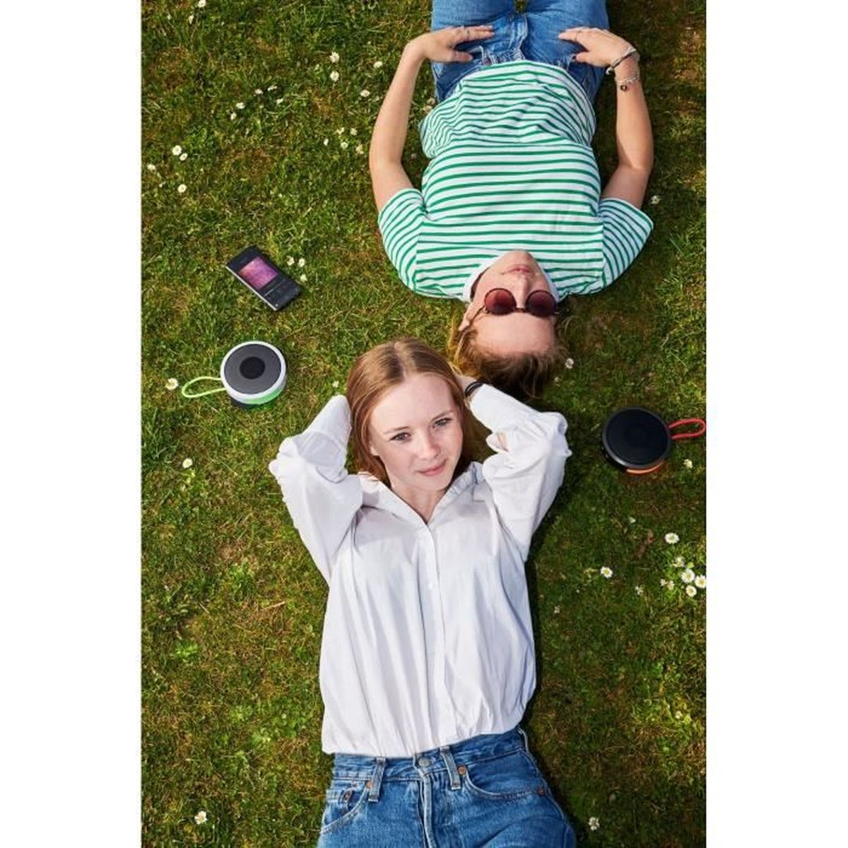 Dwie osoby leżą na trawie, patrząc w kamerę. Obok nich głośniki i telefon.