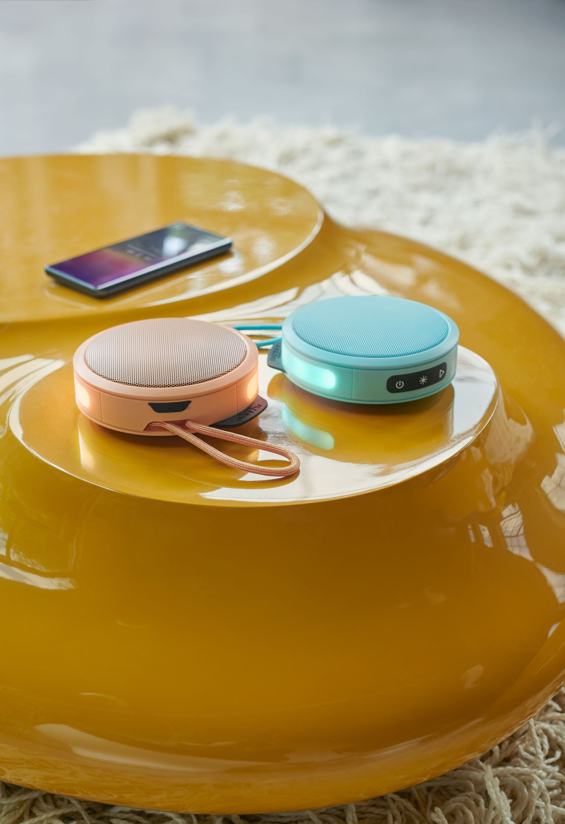 Dwa małe głośniki na żółtym stole. Jeden różowy, jeden niebieski, z telefonem.