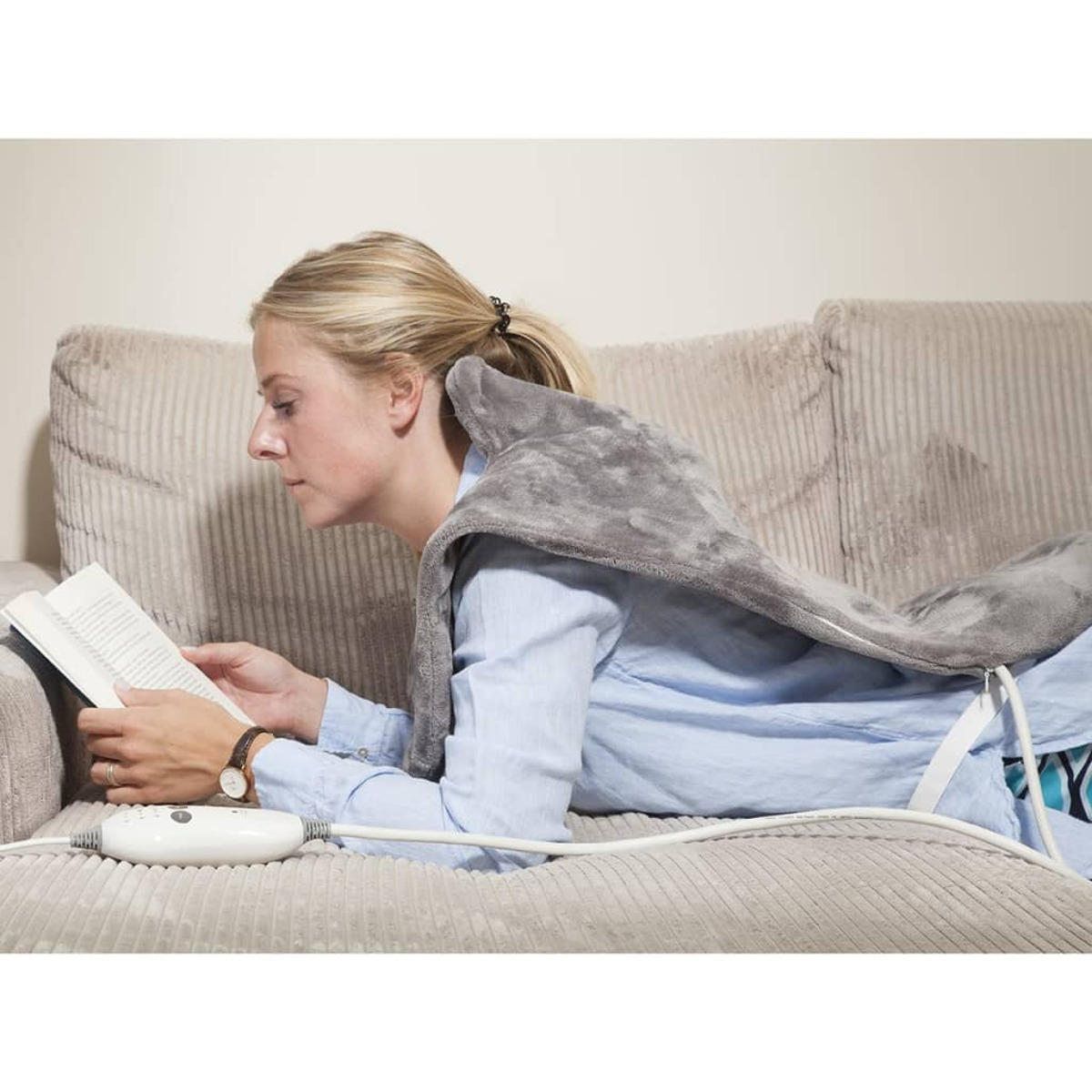 Donna che legge un libro sul divano, con un cuscinetto riscaldante grigio, con pannello di controllo.