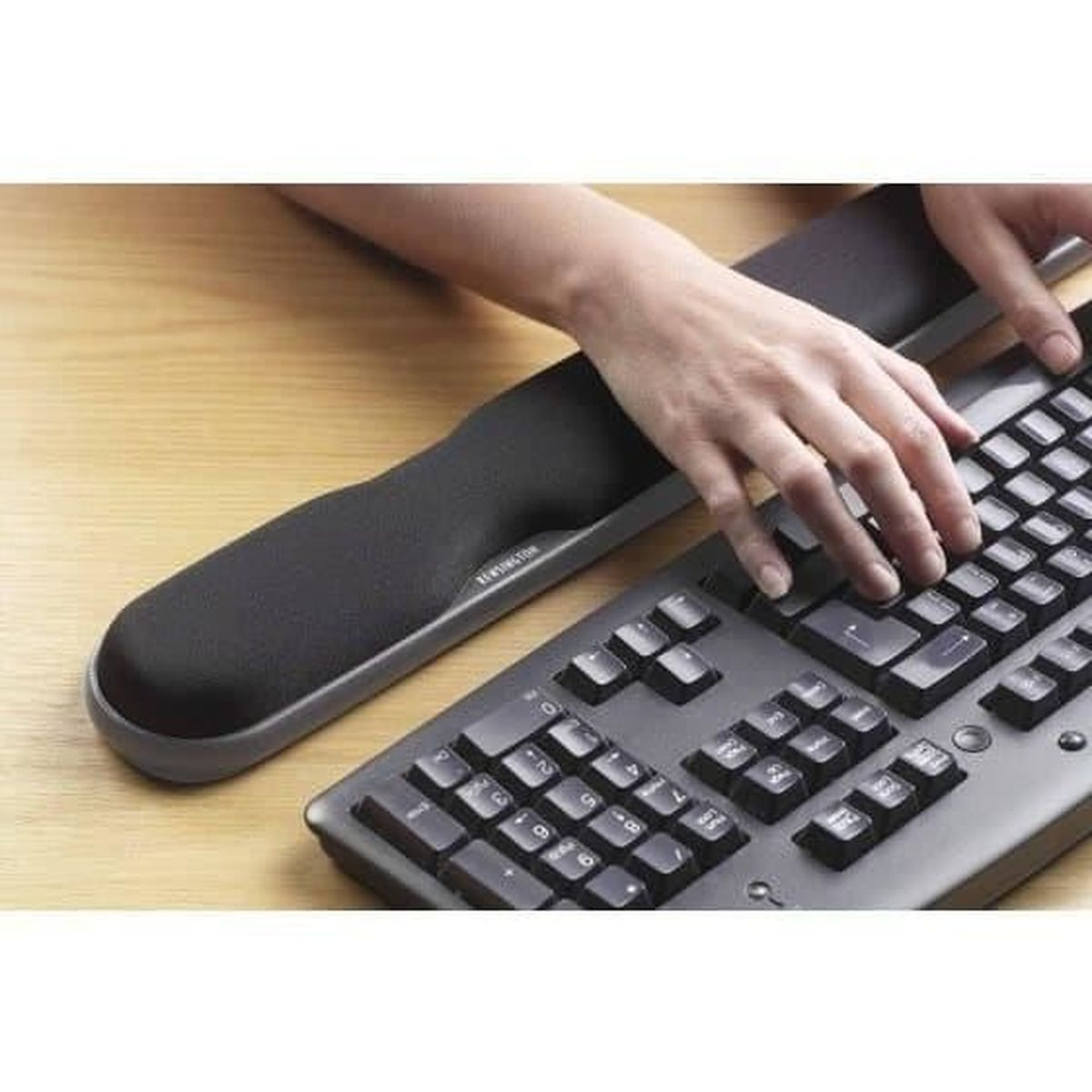 Manos tecleando en un teclado con reposamuñecas negro sobre escritorio de madera.