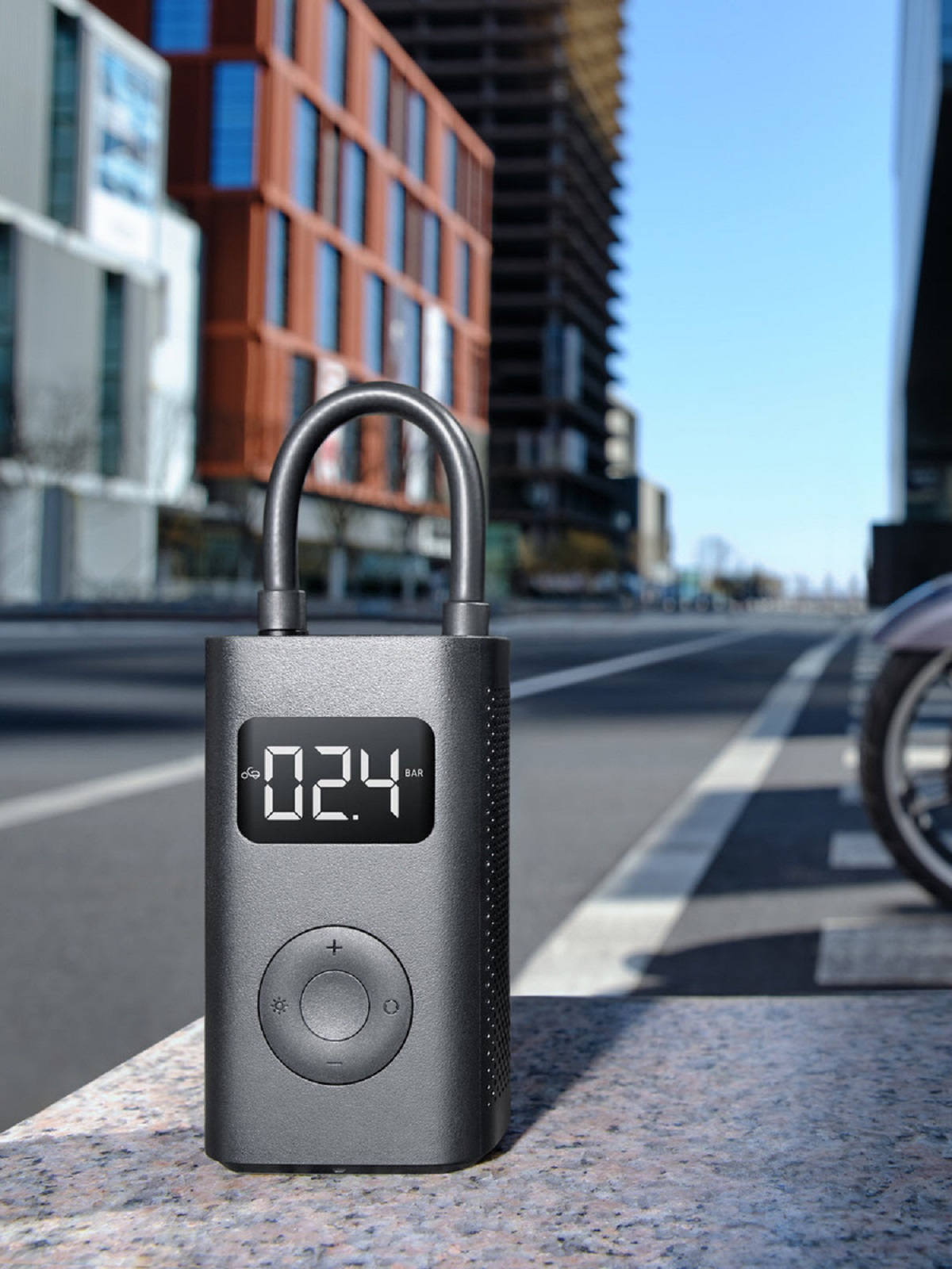 Inflador de neumáticos gris con pantalla digital que muestra 2.4 BAR. En un entorno urbano con edificios.
