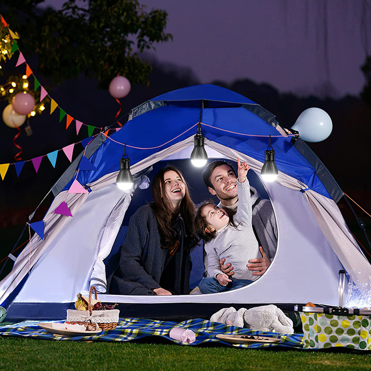 Familie im Zelt bei Nacht, mit Lichtern, Luftballons und einem Picknickkorb.