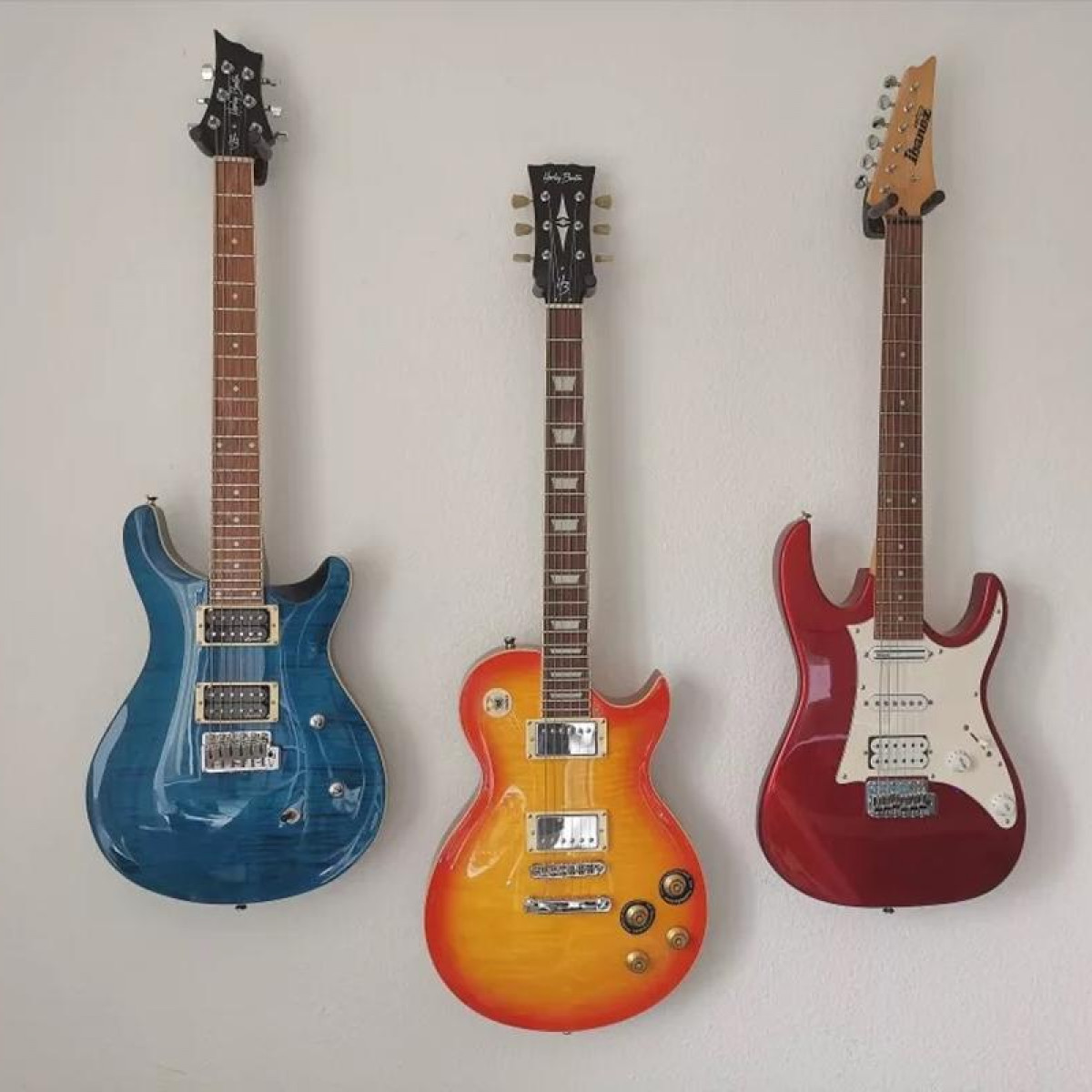 Drie gitaren hangen aan een witte muur. Een blauwe, een oranje en een rode, elk met een kop.