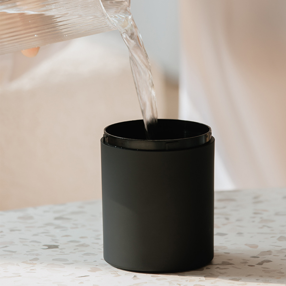 Wasser wird in eine schwarze Tasse gegossen. Die Tasse steht auf einer weißen Oberfläche. Die Szene ist drinnen.