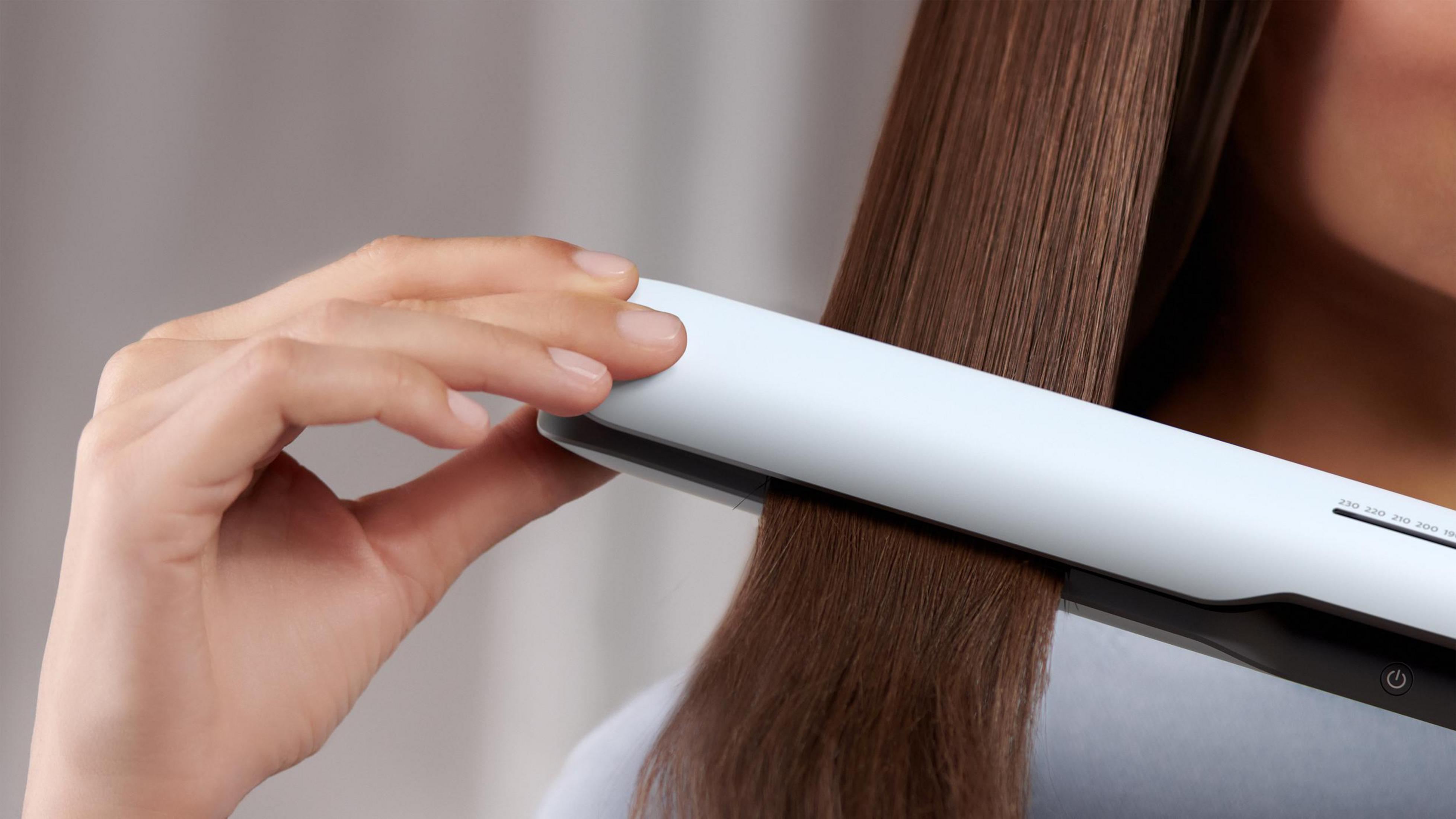 Dłoń używająca białej i srebrnej prostownicy, prostująca brązowe włosy.