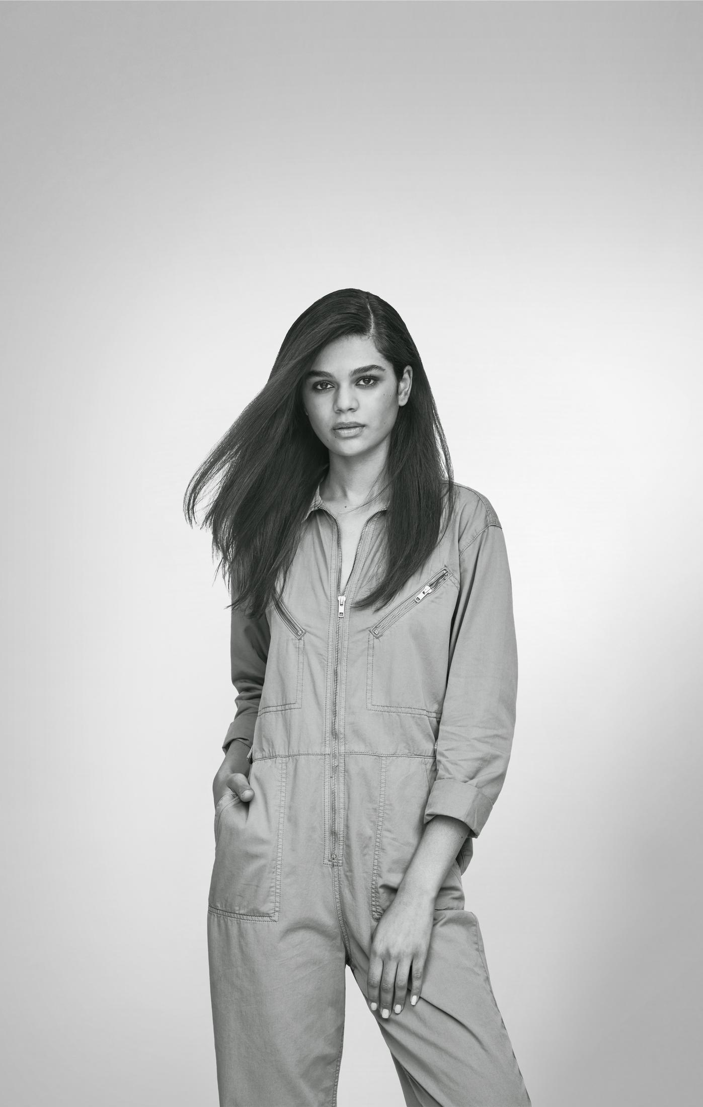 Kobieta w jasnozielonym kombinezonie, ręce w kieszeniach, patrząca przed siebie, z wiatrem we włosach.