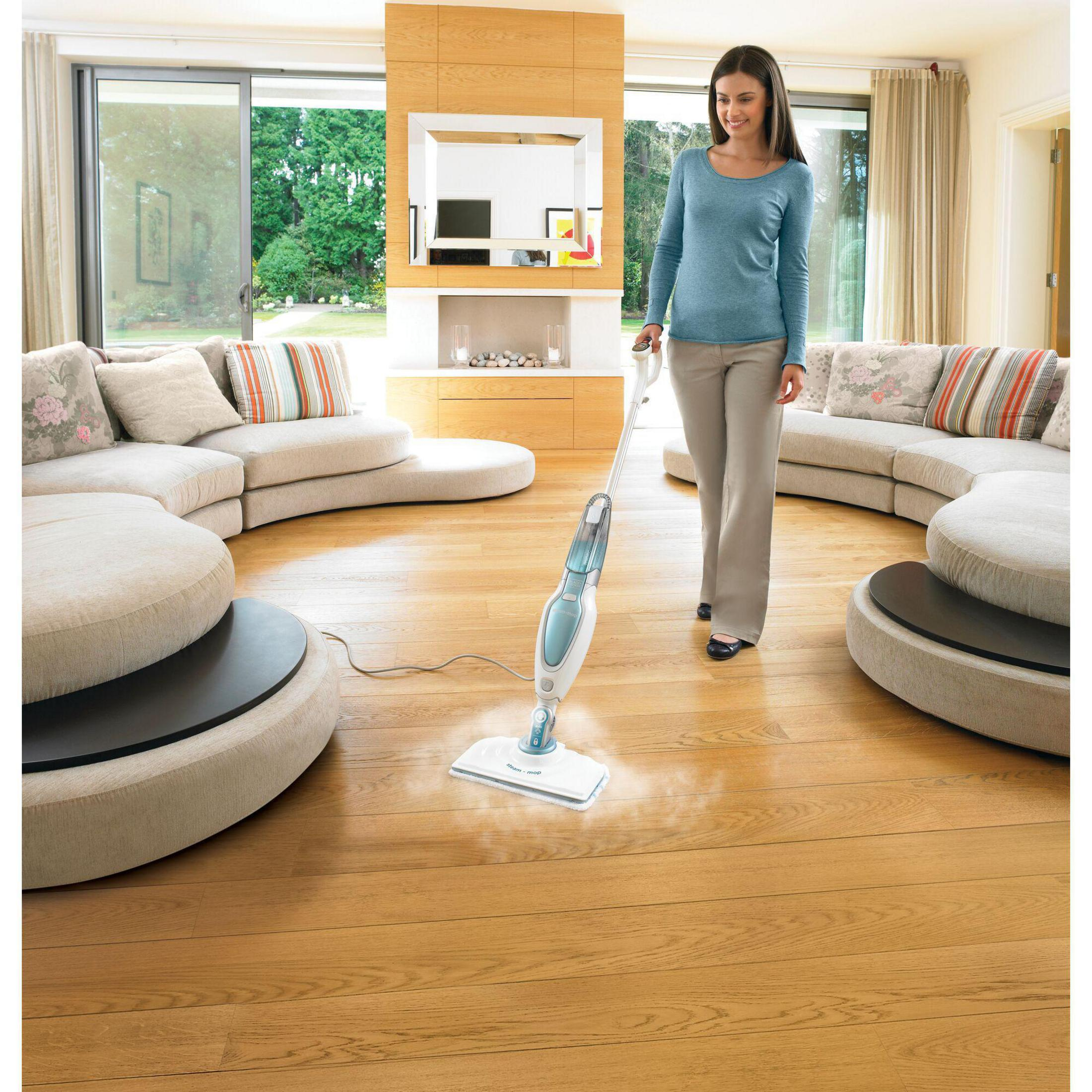 Kobieta czyści drewnianą podłogę mopem parowym w nowoczesnym salonie z okrągłymi sofami. Światło słoneczne.