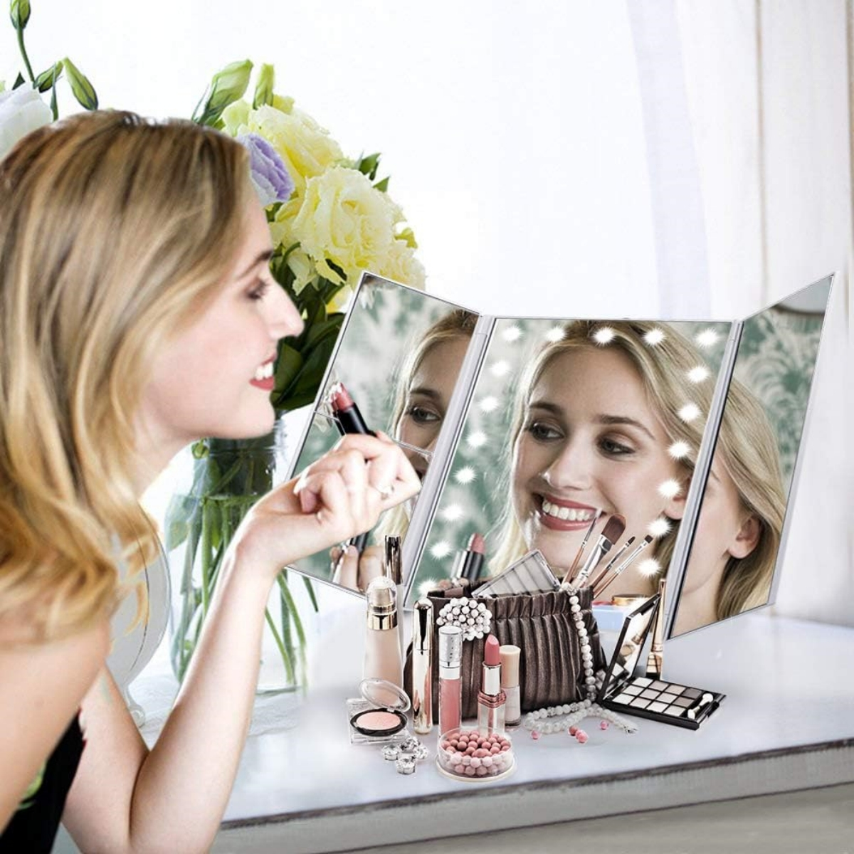 Eine Frau, die sich in einem dreiteiligen Spiegel Lippenstift aufträgt. Schminkartikel und Blumen stehen auf dem Tisch.
