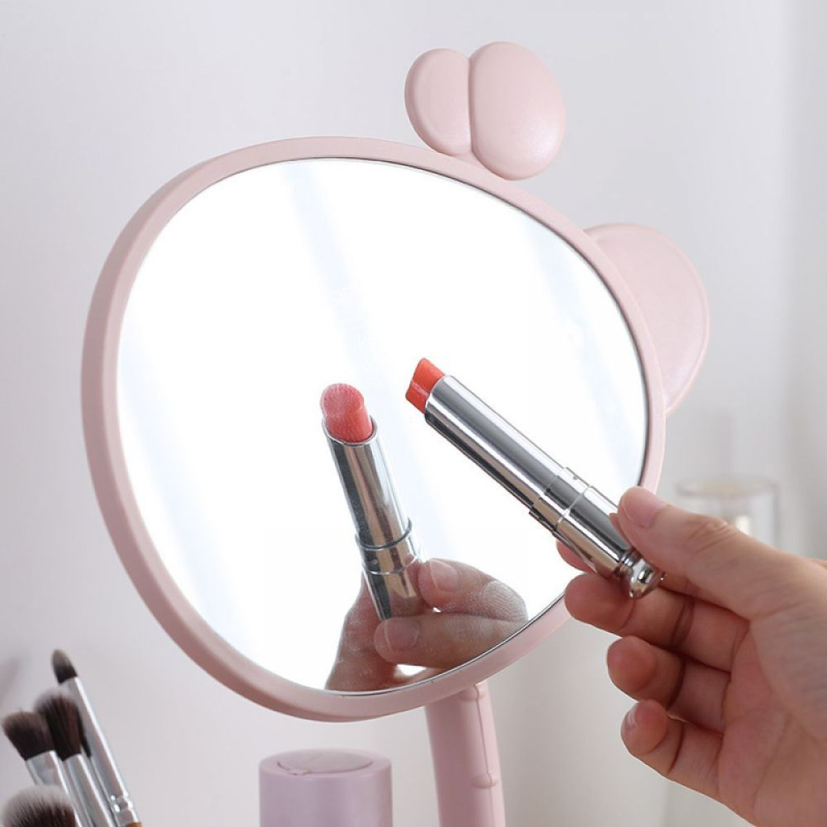Eine Frau hält einen Lippenstift neben einem rosa, runden Spiegel.