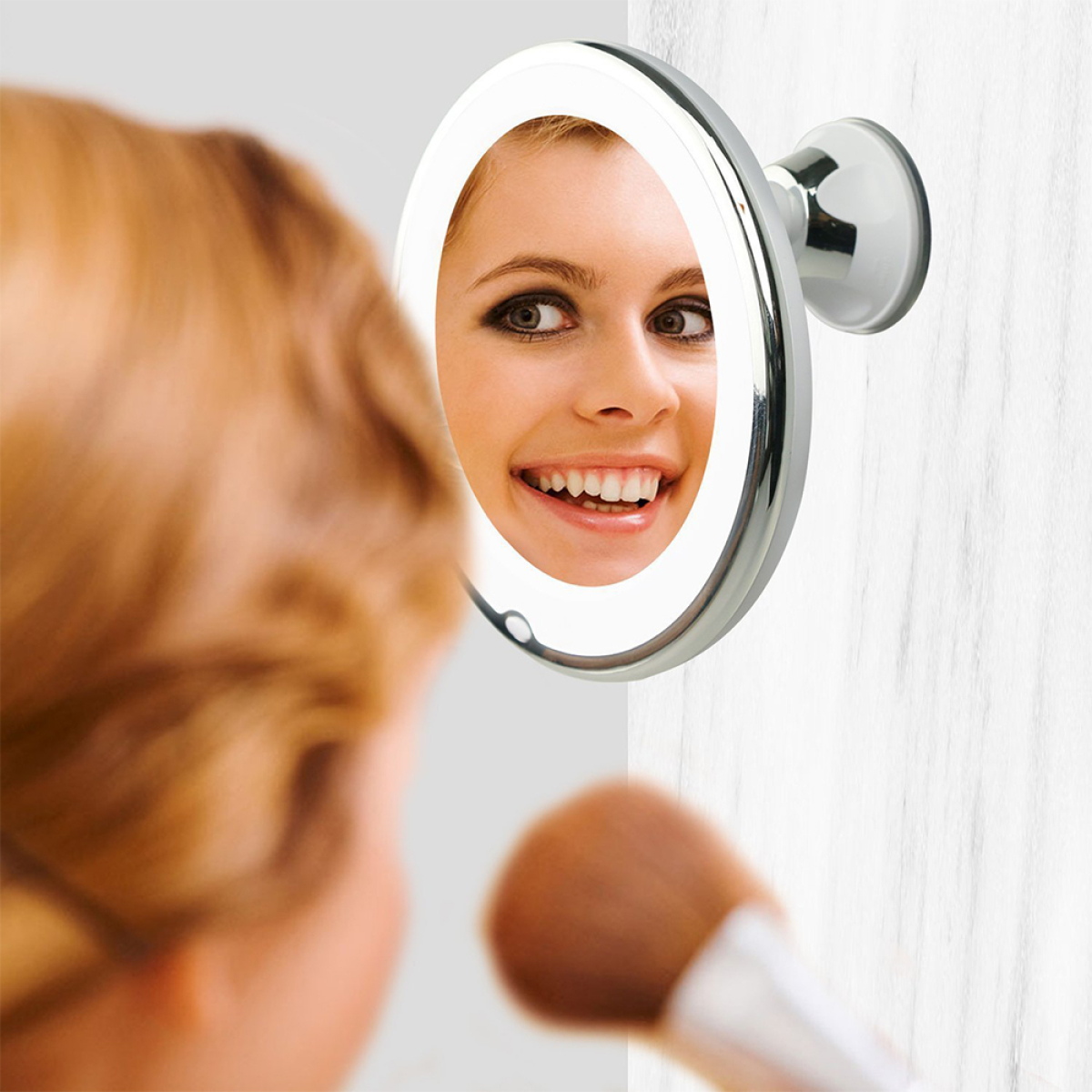 Frau schaut in beleuchteten, runden Spiegel, trägt Make-up mit Pinsel auf. Weiße Wand.