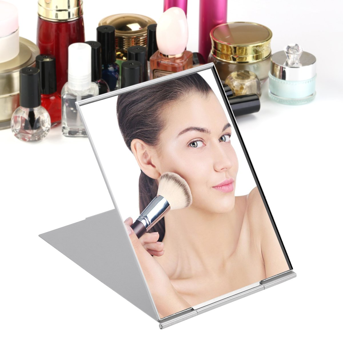 Eine Frau trägt Make-up mit einem Pinsel vor einem silbernen, faltbaren Spiegel auf, umgeben von Kosmetika.