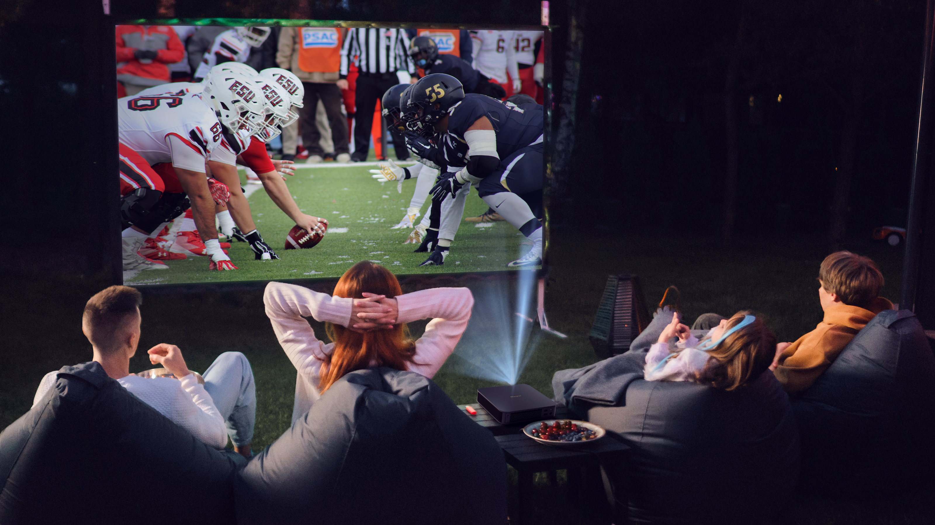 Ludzie oglądający wyświetlany mecz futbolu na ekranie zewnętrznym, siedząc na workach sako.