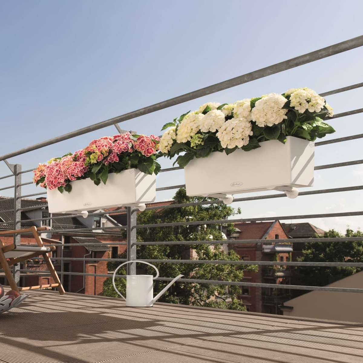 Zwei weiße Blumenkästen mit rosa und weißen Blumen auf einem Balkongeländer, sonniger Himmel im Hintergrund.