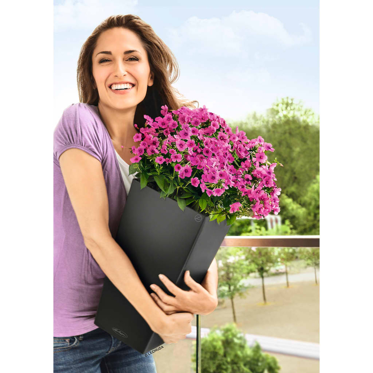 Frau hält einen schwarzen Pflanzkübel mit rosa Blumen auf einem Balkon. Himmel im Hintergrund.