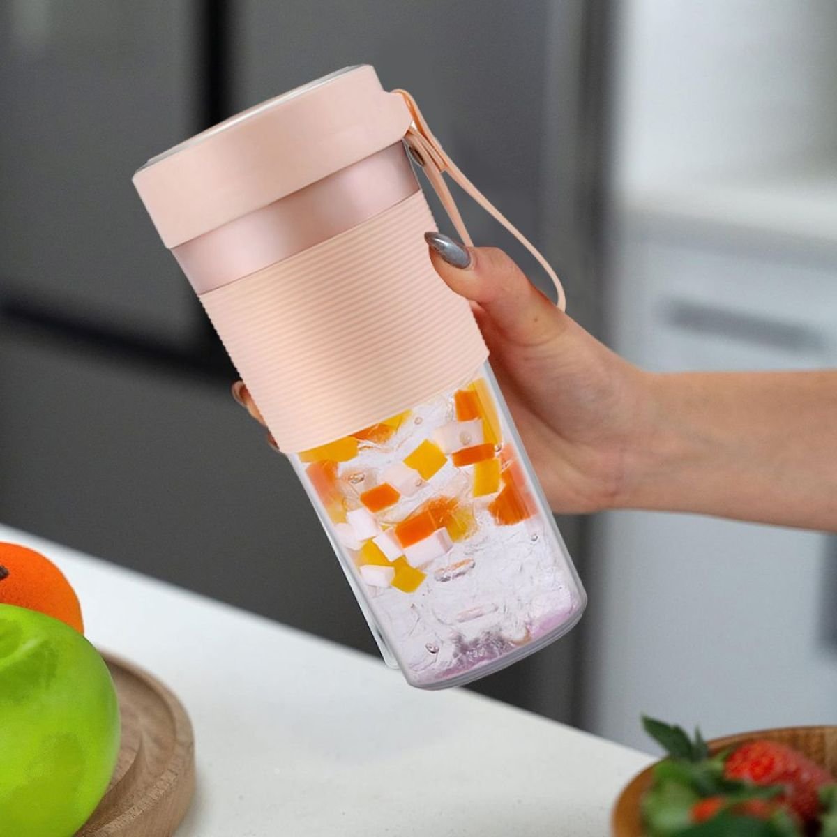 Eine Hand hält einen Mixer mit Fruchtwürfeln und Eis. Ein grüner Apfel und Erdbeeren sind im Vordergrund.