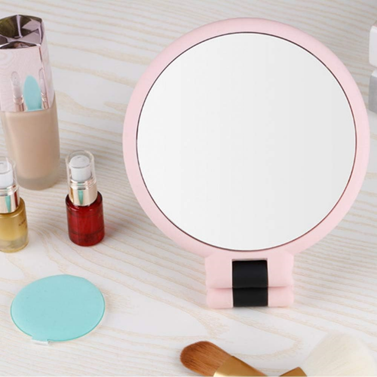 Ein rosa Spiegel, kleine Make-up-Fläschchen und ein Make-up-Pinsel liegen auf einem Holztisch.
