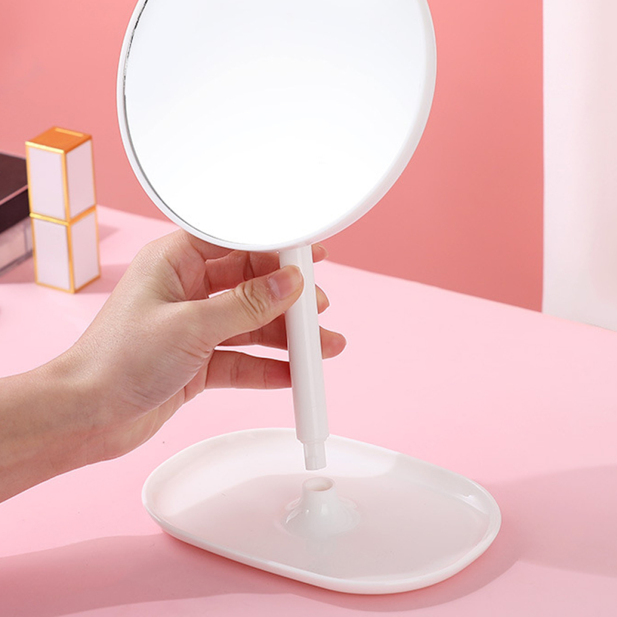 Ein weißer Spiegel mit Ständer, Hand, die ihn hält, auf einer rosa Oberfläche mit Objekten.