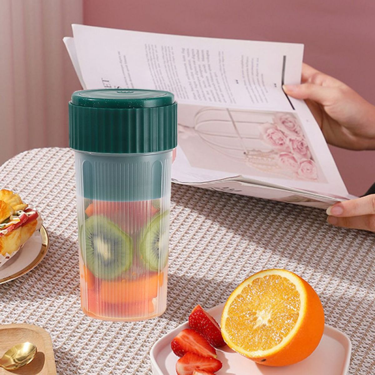 Tragbarer Mixer mit Früchten, neben Orange, Erdbeeren und einem Buch.