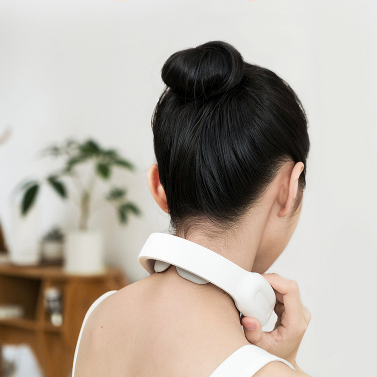 Eine Frau trägt ein weißes Nackenmassagegerät. Ihre Haare sind zu einem Dutt gebunden. Rückansicht.