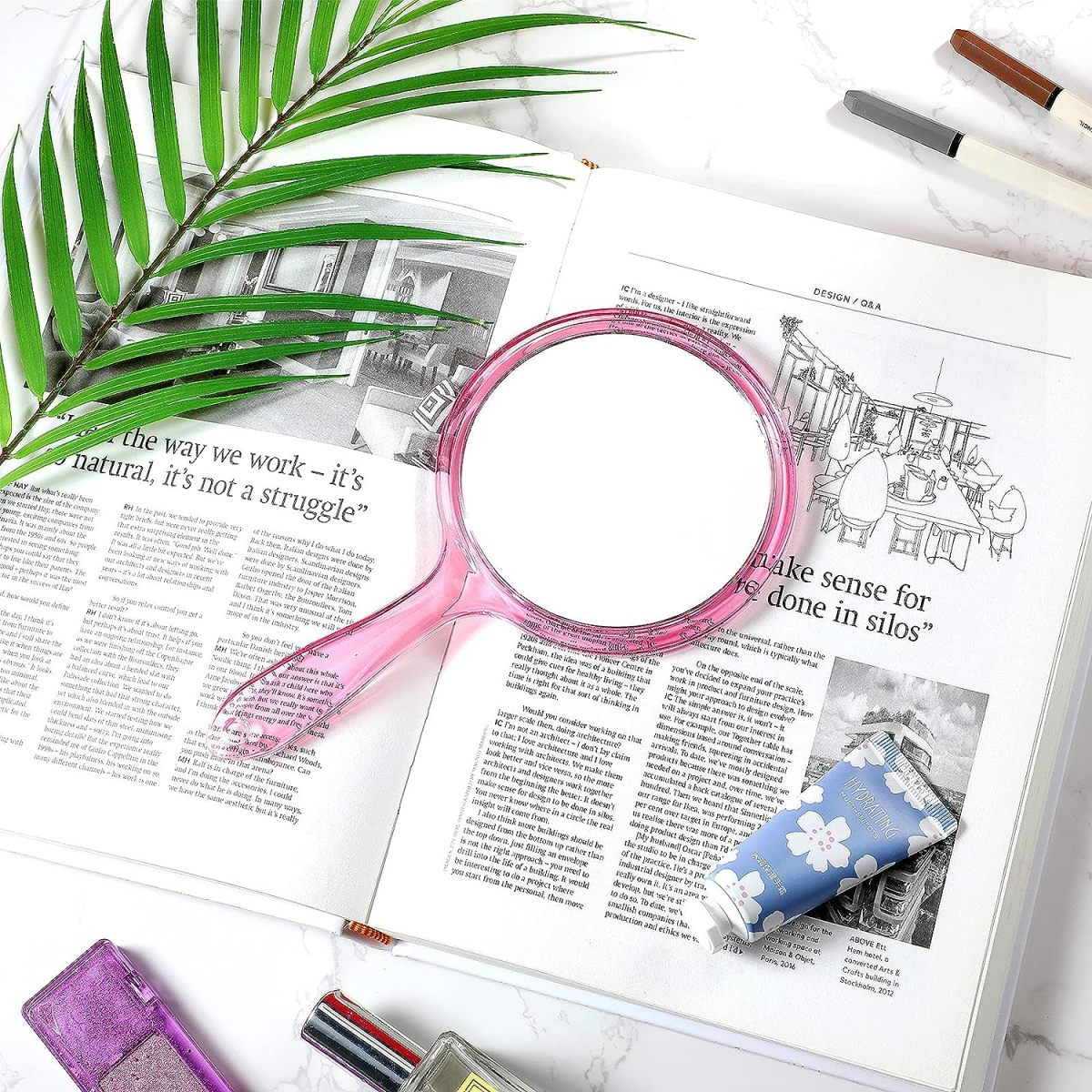 Ein Handspiegel auf einer offenen Zeitschrift mit Make-up und einer Pflanze. Der Spiegel hat einen rosa Griff.