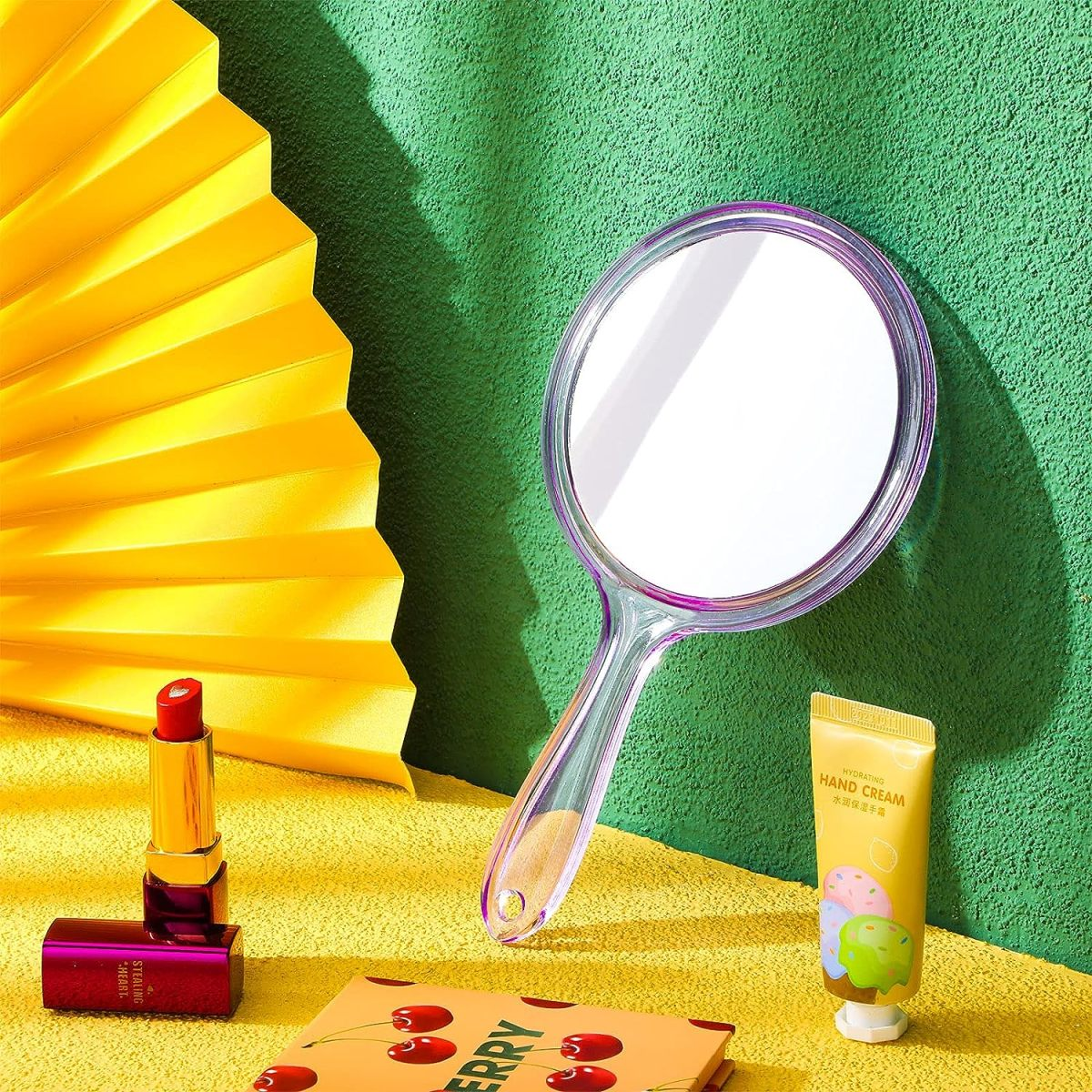 Ein Spiegel, Lippenstift, Handcreme und ein Buch auf gelbem Untergrund mit grünem Hintergrund.