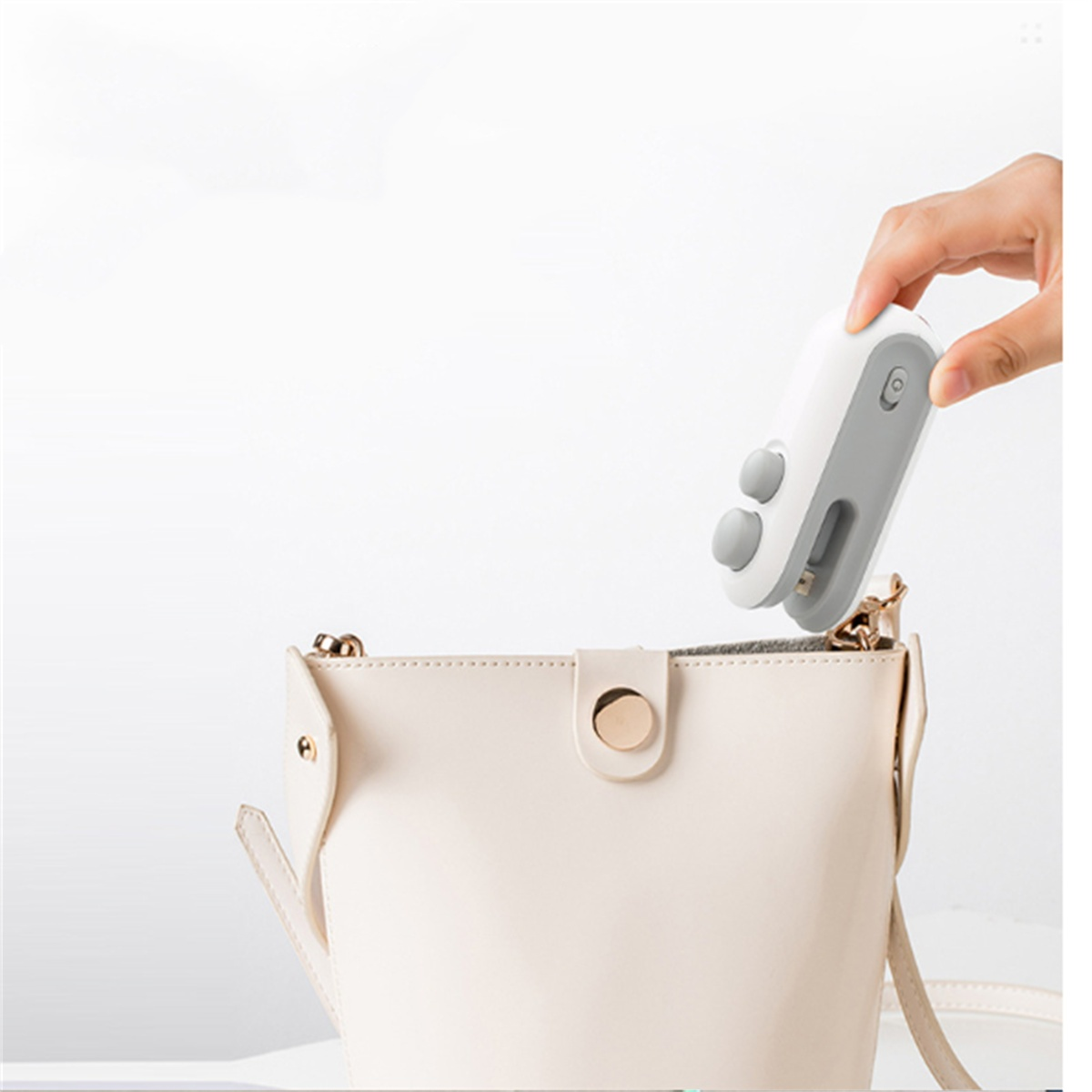 Hand steckt ein weiß-graues Gerät in eine hellbeige Handtasche. Weißer Hintergrund.
