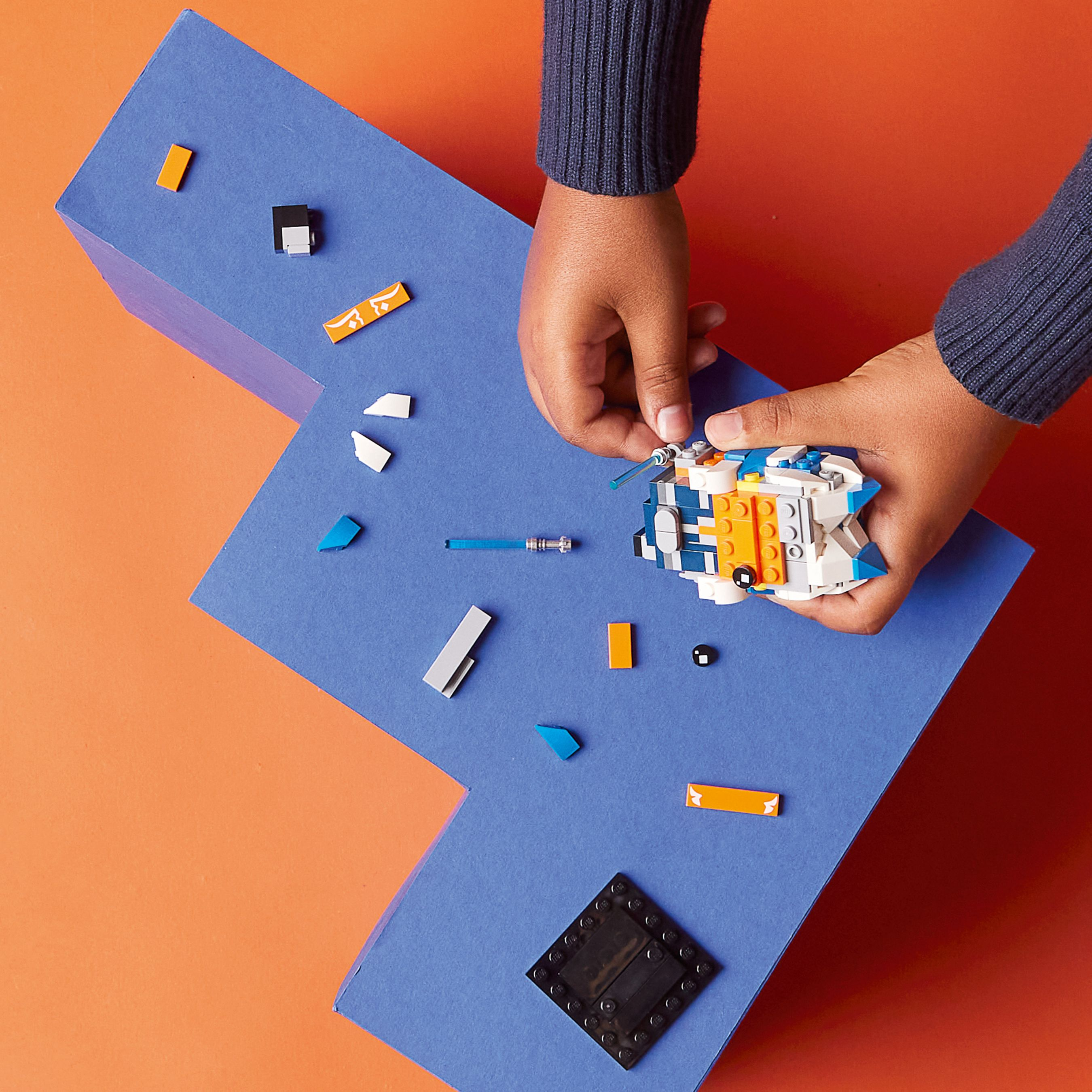 Hände, die ein Lego-Spielzeug auf blauem Papier vor einem orangefarbenen Hintergrund zusammensetzen. Legosteine ​​liegen herum.
